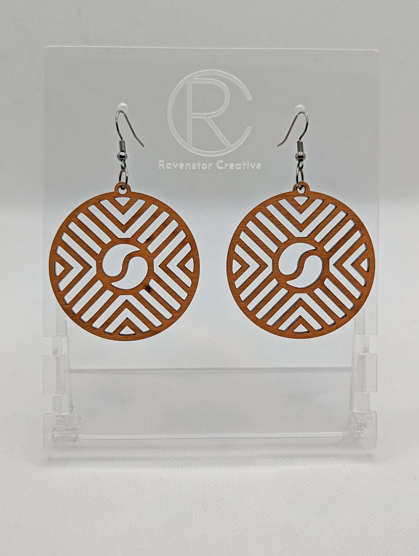 Shows a pair of Circle earrings that have black ear wire fixings. They are Geometric Chevron designs with a yin and yang in the centre. They are made from Cherry Wood. They are against a white background on see through stands.