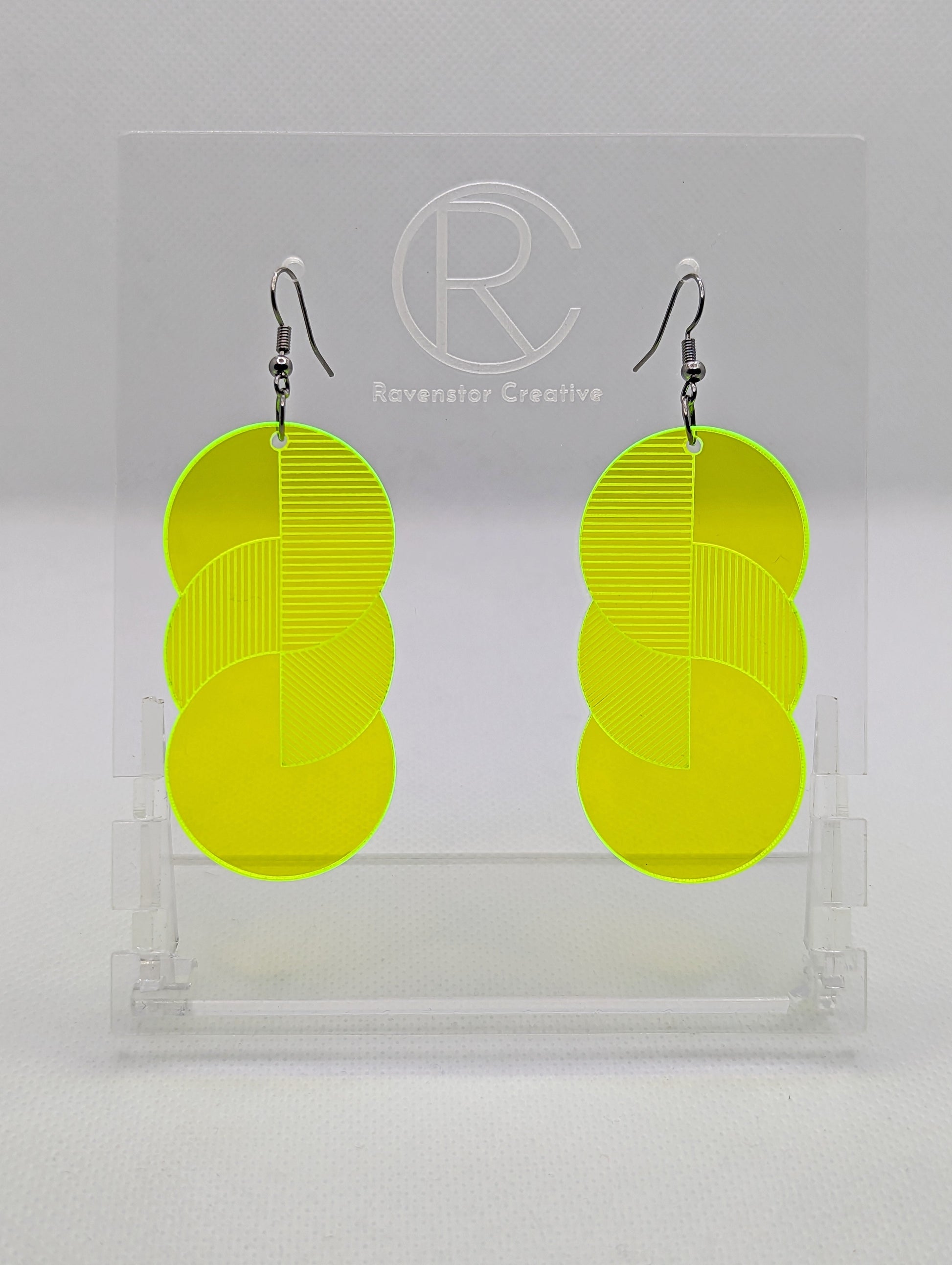 A pair of large acrylic earrings with black ear wire hook fixings. Each earring is 3 circles overlapping and entwined with  engraved line decorations. The colour is neon Green and they are hung on a clear stand against a white background.