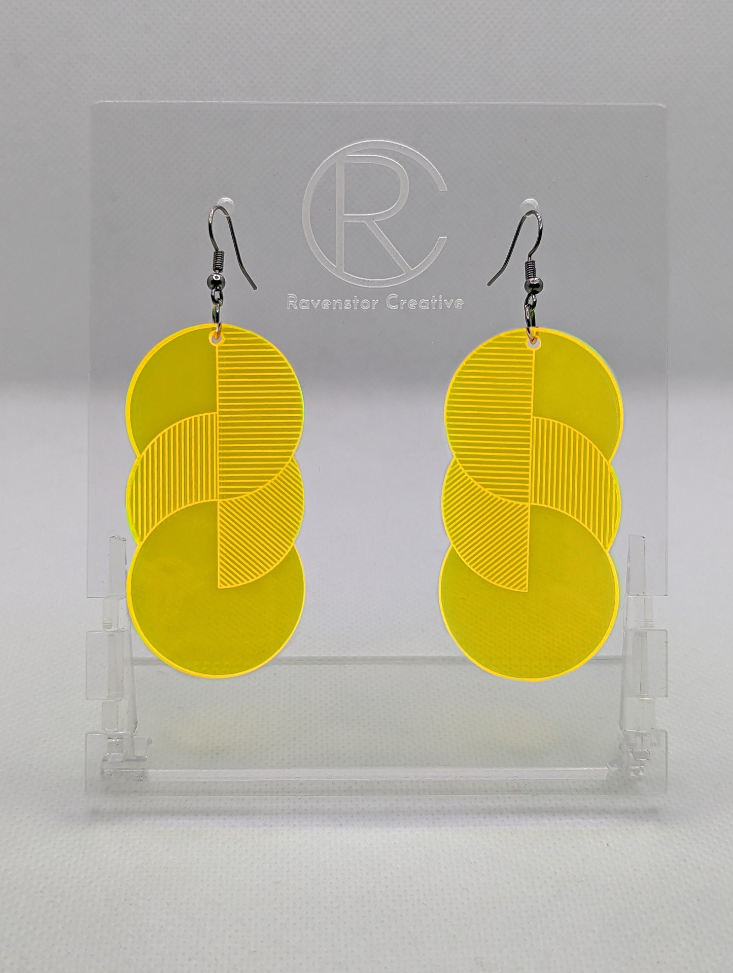 A pair of large acrylic earrings with black ear wire hook fixings. Each earring is 3 circles overlapping and entwined with  engraved line decorations. The colour is neon orange and they are hung on a clear stand against a white background.