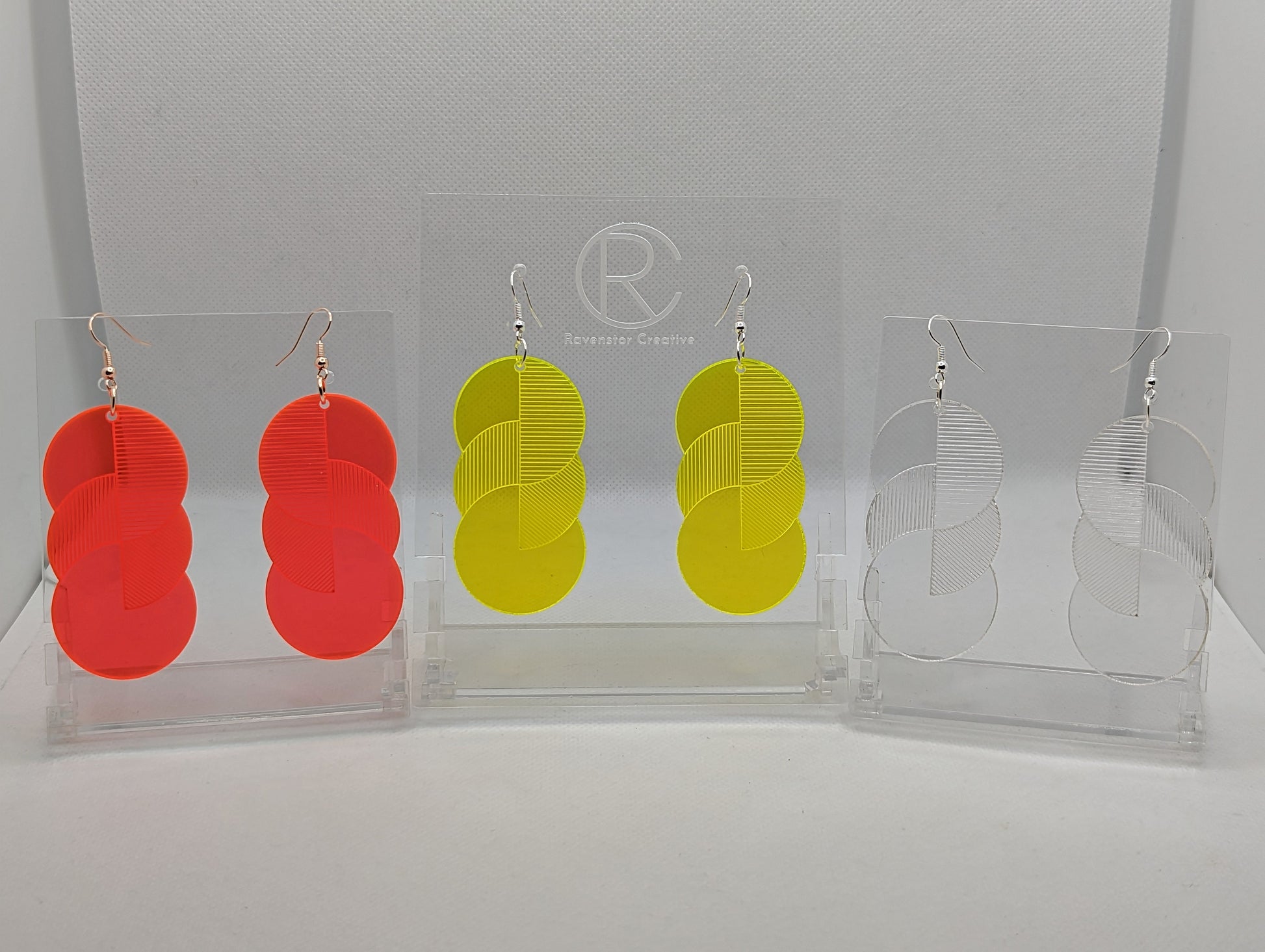 Three pairs of large acrylic earrings. Each one is 3 circles overlapping and entwined with  engraved line decorations hung on a clear stand. The colours are Neon pink, Neon Yellow and clear / See through. They have silver ear wire hook fixings.