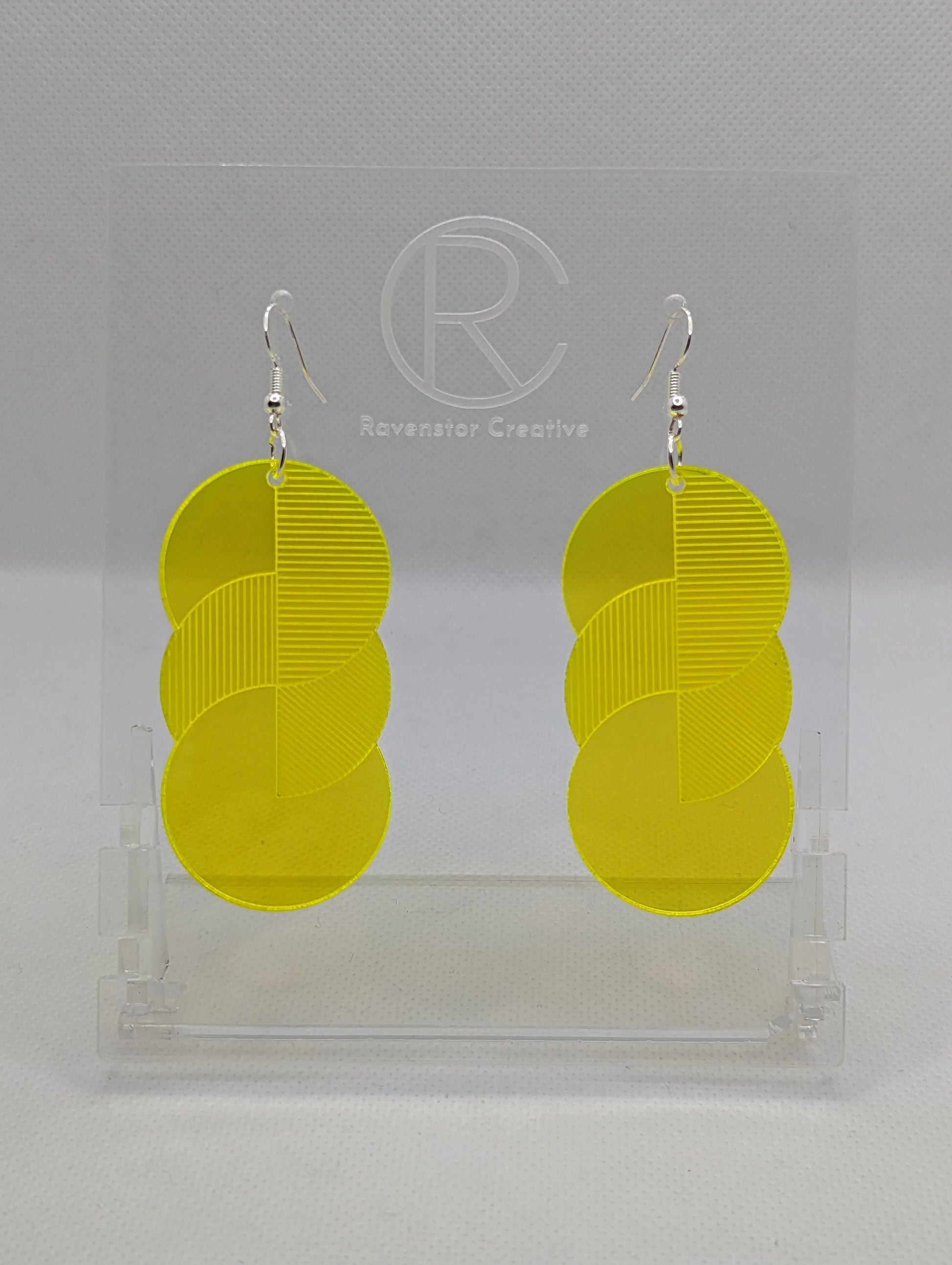 A pair of large acrylic earrings with silver ear wire hook fixings. Each earring is 3 circles overlapping and entwined with  engraved line decorations. The colour is neon yellow and they are hung on a clear stand against a white background.