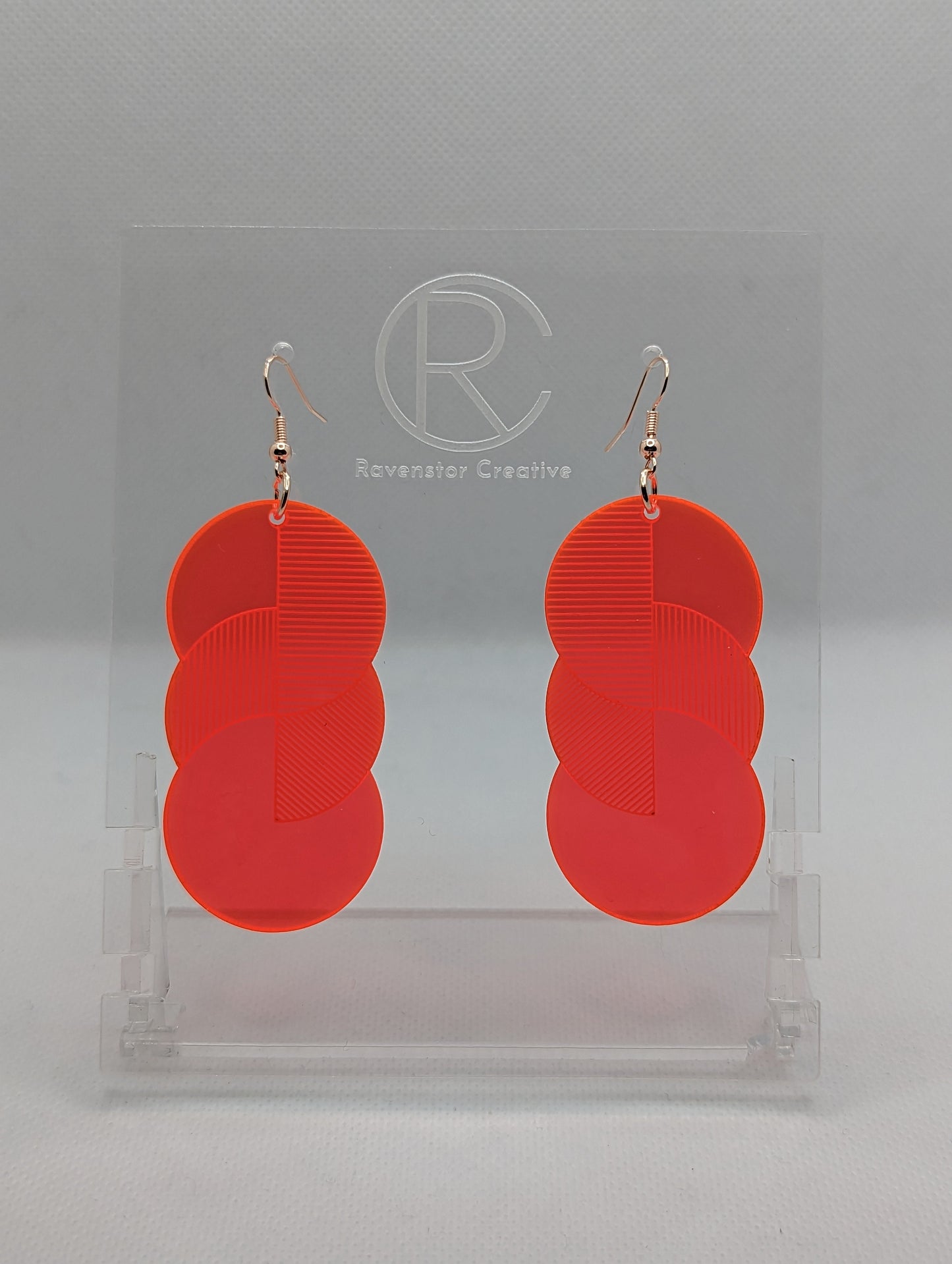 A pair of large acrylic earrings with silver ear wire hook fixings. Each earring is 3 circles overlapping and entwined with  engraved line decorations. The colour is neon pink and they are hung on a clear stand against a white background.