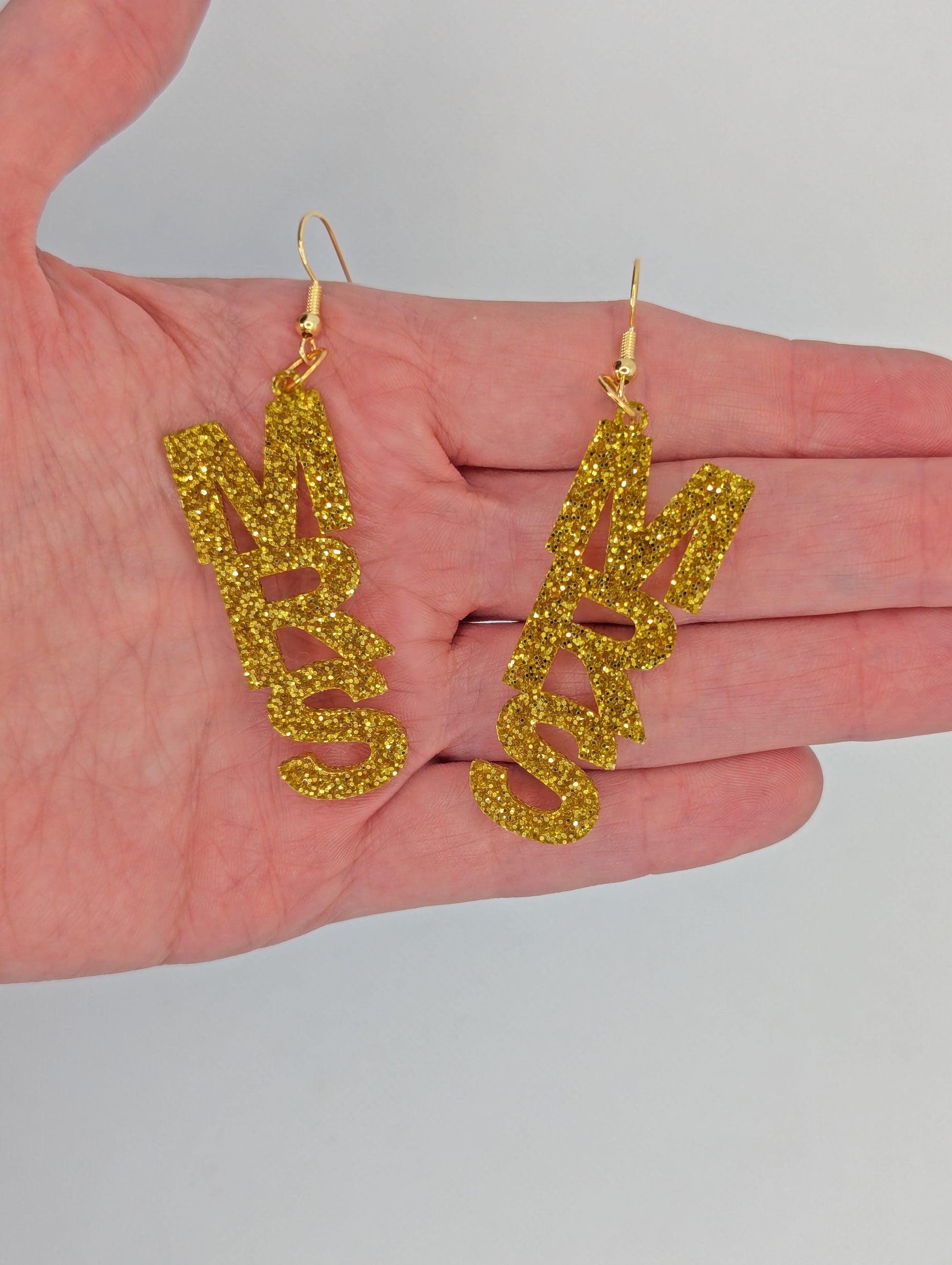 Earrings with Gold ear wire hook fixings. Each one is the word Mrs in capital letters. These are ideal for a Hen do or Bachelorette Party. They are against a hand to show the size of three finger widths in length and one and a half wide.