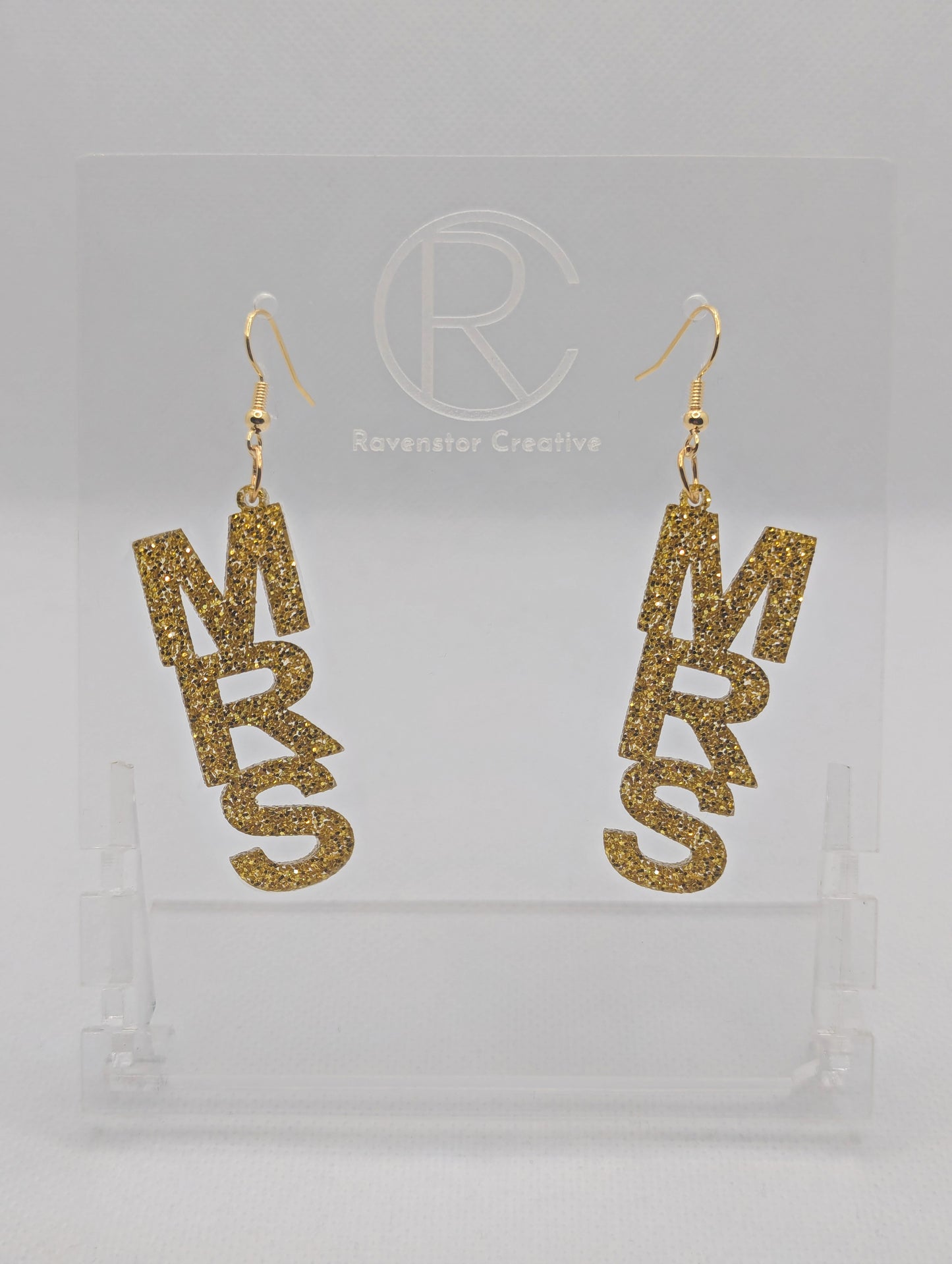 A pair of earrings with Gold ear wire hook fixings. Each one is the word Mrs in capital letters. They are Gold Glitter in colour. These are ideal for a Hen do or Bachelorette Party. They are on clear stands against a white background.