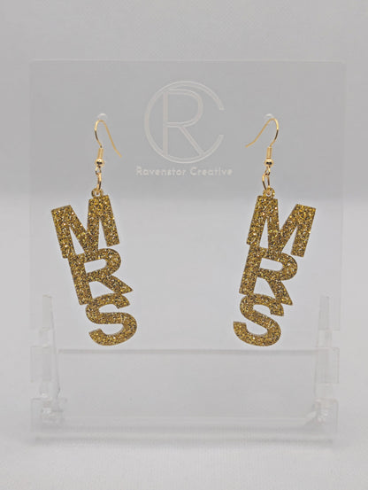 A pair of earrings with Gold ear wire hook fixings. Each one is the word Mrs in capital letters. They are Gold Glitter in colour. These are ideal for a Hen do or Bachelorette Party. They are on clear stands against a white background.