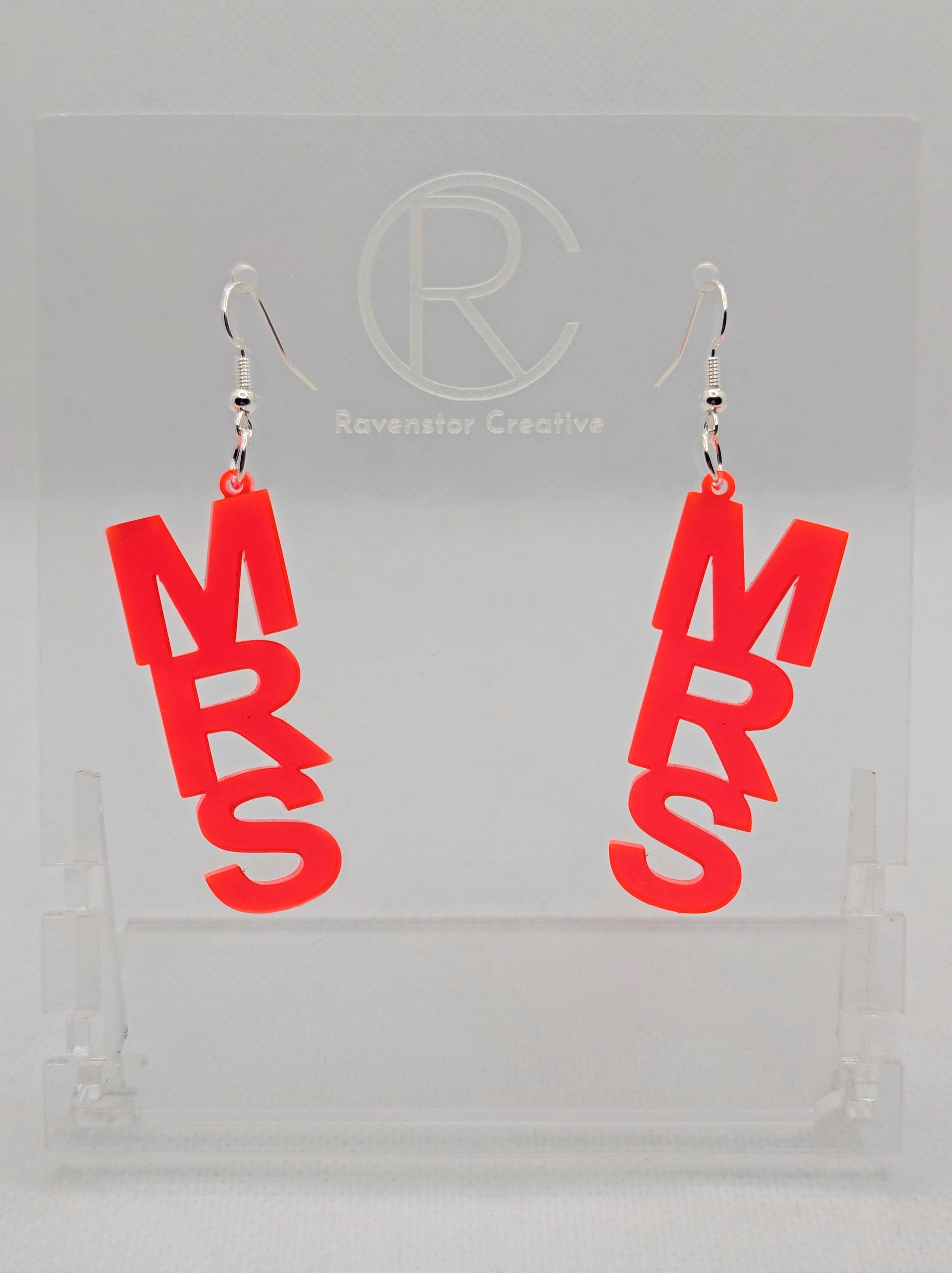 A pair of earrings with Silver ear wire hook fixings. Each one is the word Mrs in capital letters. They are Neon Pink in colour. These are ideal for a Hen do or Bachelorette Party. They are on clear stands against a white background.