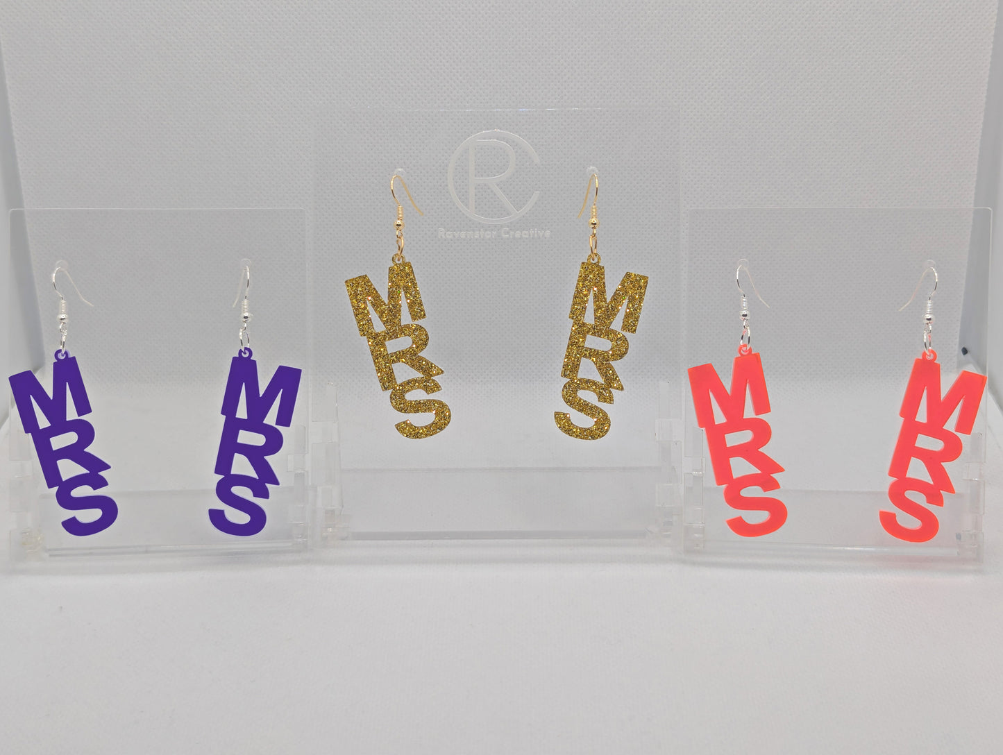 Image of Three pairs of earrings with ear wire hook fixings. Each one is the word Mrs in capitals. The colours are Purple, Gold Glitter and Sherbet Pink. They are on clear stands against a white background.