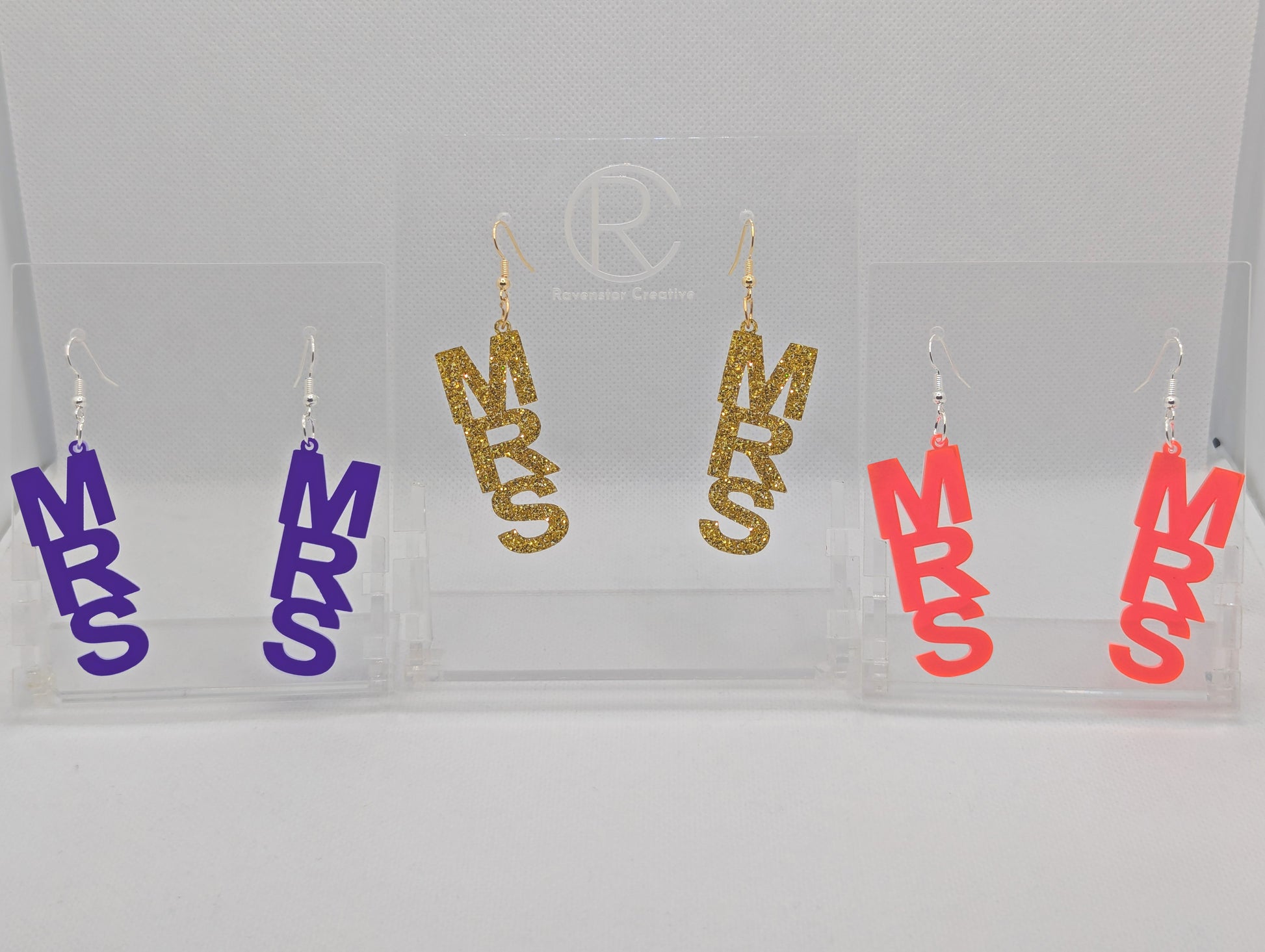 Image of Three pairs of earrings with ear wire hook fixings. Each one is the word Mrs in capitals. The colours are Purple, Gold Glitter and Sherbet Pink. They are on clear stands against a white background.