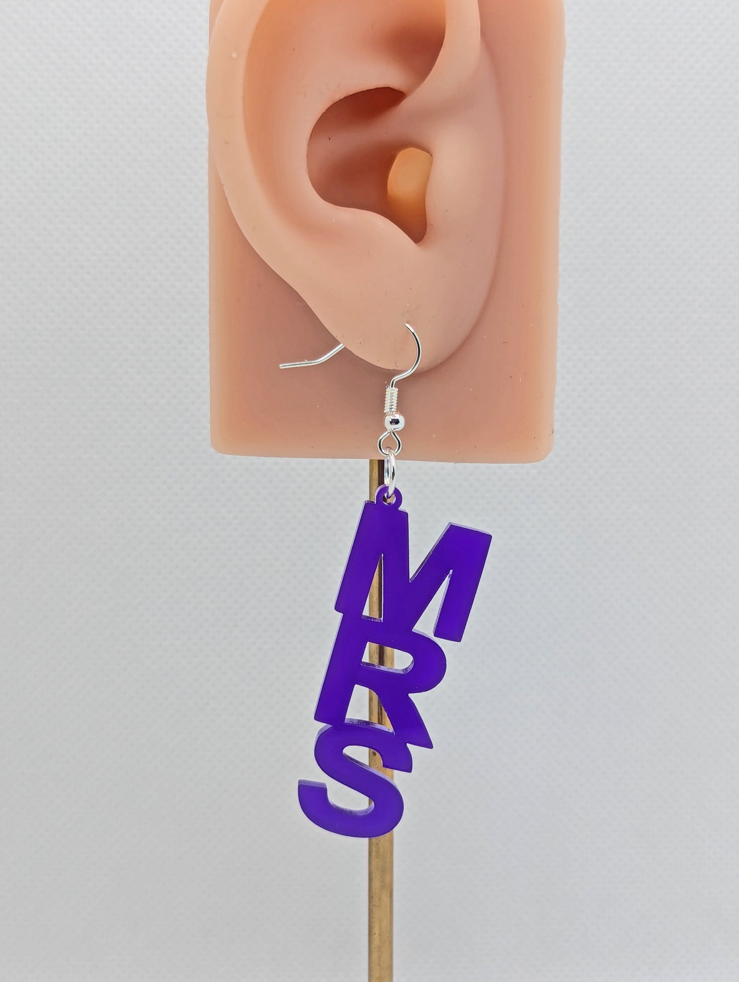 An earring with a Silver ear wire hook fixing. It is the word Mrs in capital letters. It is Purple in colour. These are ideal for a Hen do or Bachelorette Party. They are on an ear to show the size.