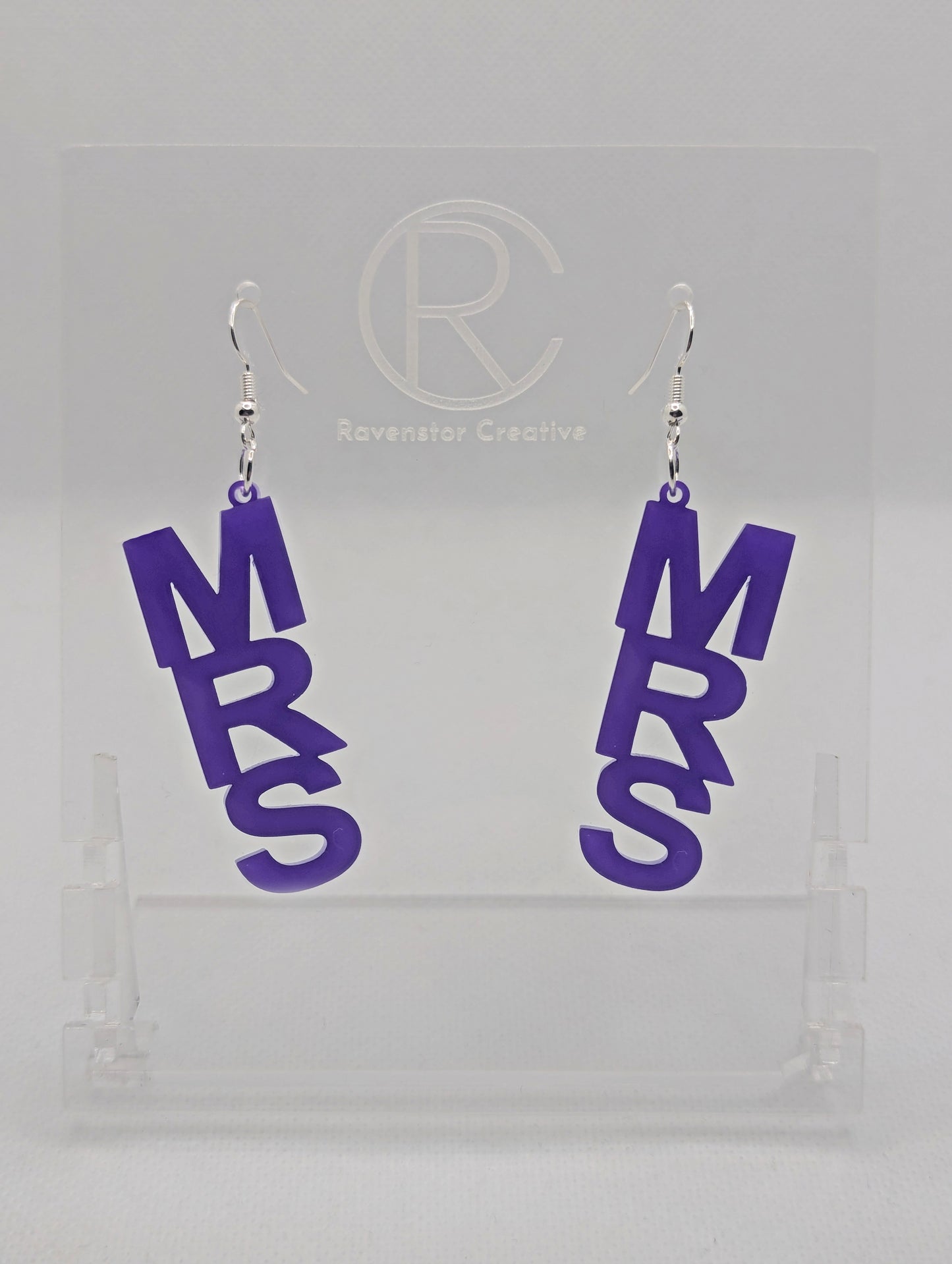 A pair of earrings with Silver ear wire hook fixings. Each one is the word Mrs in capital letters. They are Purple in colour. These are ideal for a Hen do or Bachelorette Party. They are on clear stands against a white background.