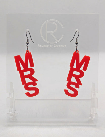 A pair of earrings with Black ear wire hook fixings. Each one is the word Mrs in capital letters. They are Red in colour. These are ideal for a Hen do or Bachelorette Party. They are on clear stands against a white background.