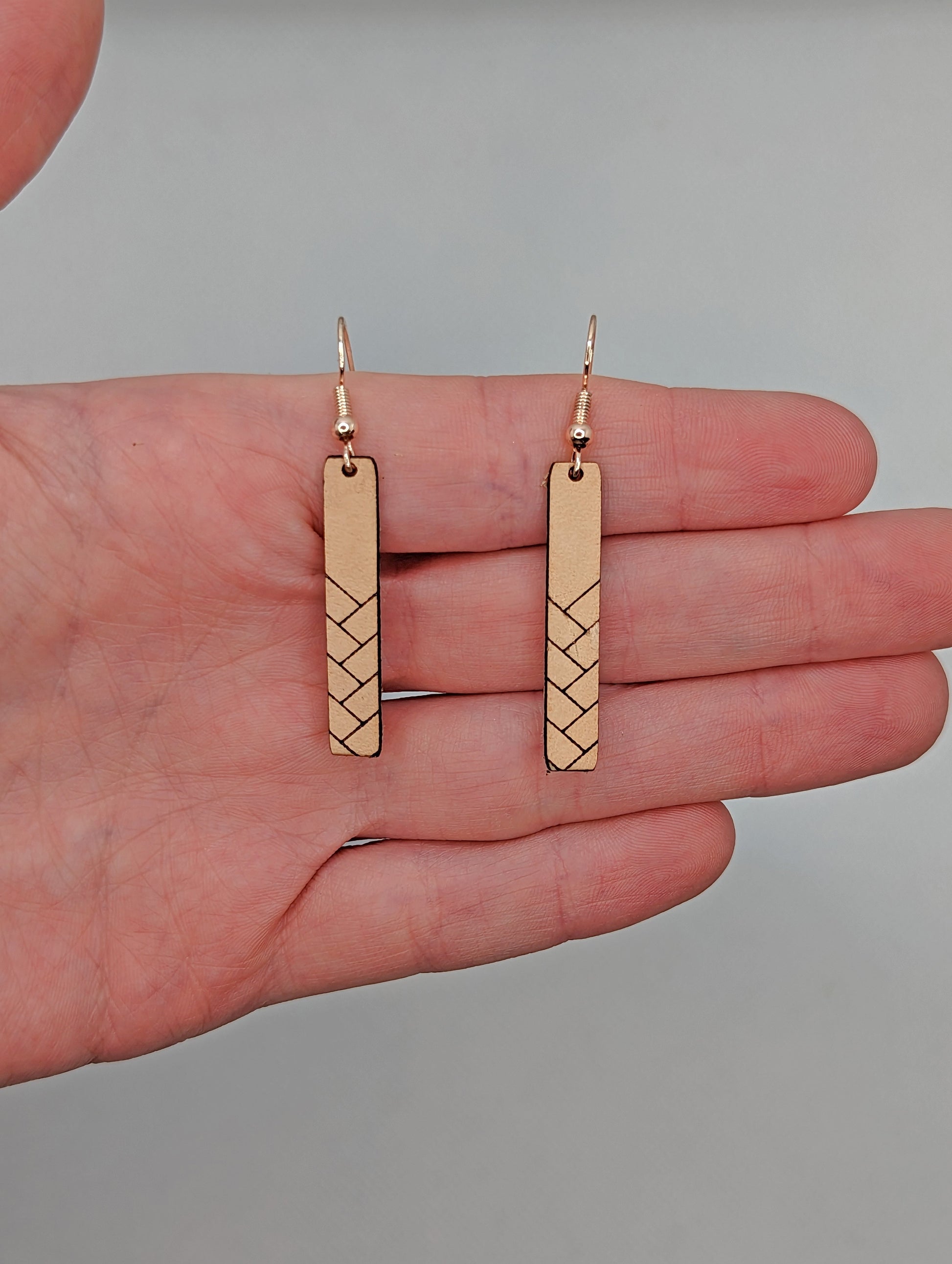 A pair or Natural Tan Leather earrings with rose gold fixings. They are thin and long and have a weave pattern They are against a hand showing the size of two and a half finger widths in length and one third finger width wide.