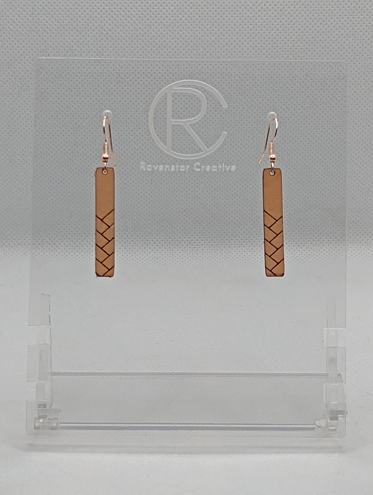 A pair or Natural Tan Leather earrings with rose gold fixings. They are thin and long and have a weave pattern that starts in the middle and goes down to the bottom. They against a white background on a clear stand with the Ravenstor Creative logo.