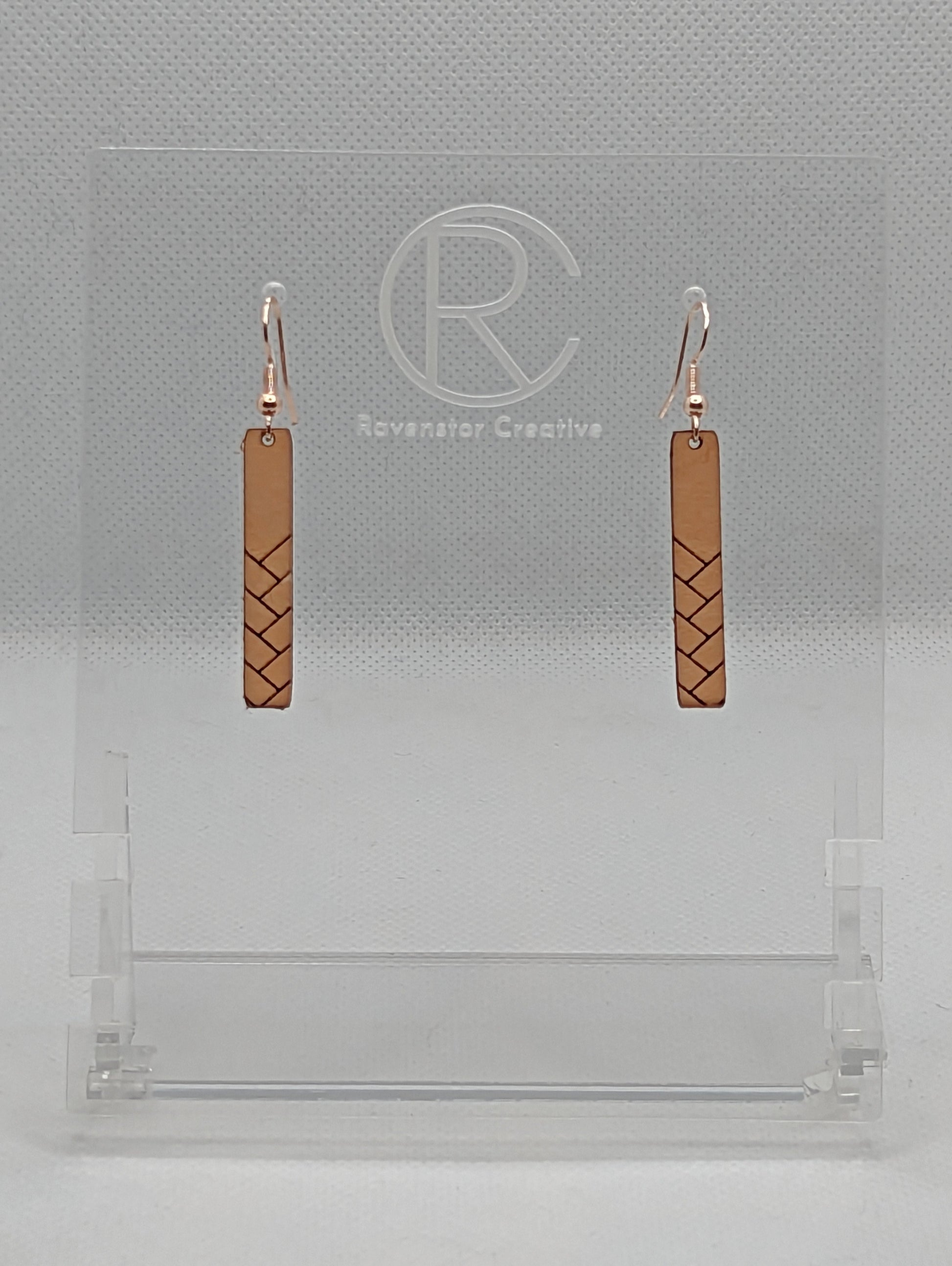 A pair or Natural Tan Leather earrings with rose gold fixings. They are thin and long and have a weave pattern that starts in the middle and goes down to the bottom. They against a white background on a clear stand with the Ravenstor Creative logo.