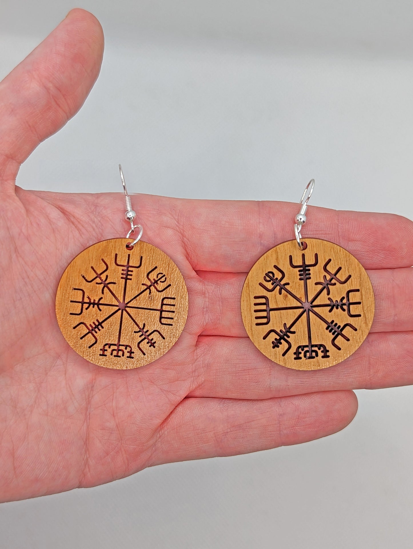 A pair of circle earrings with silver ear wire hook fixings. Cut out of the centre is the Nordic Compass charm. They are made from cherry wood. They are against a hand to show the size of two finger widths in length and same again wide