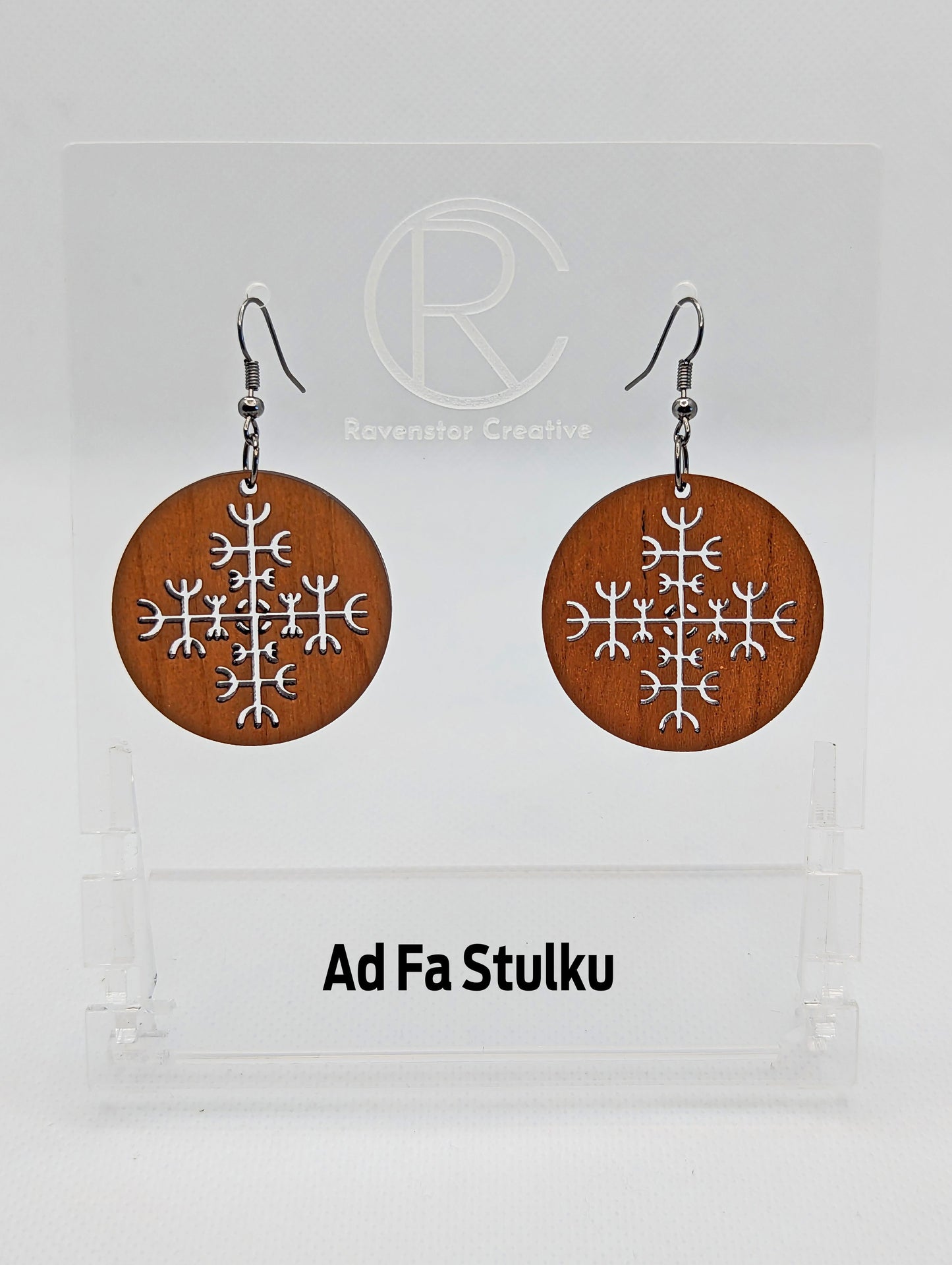 A pair of circle earrings with black ear wire hook fixings. Cut out of the centre is the Ad Fa Stulku Nordic charm. They are made from cherry wood. They are against a white background on a clear stand with the Ravenstor Creative logo on it.