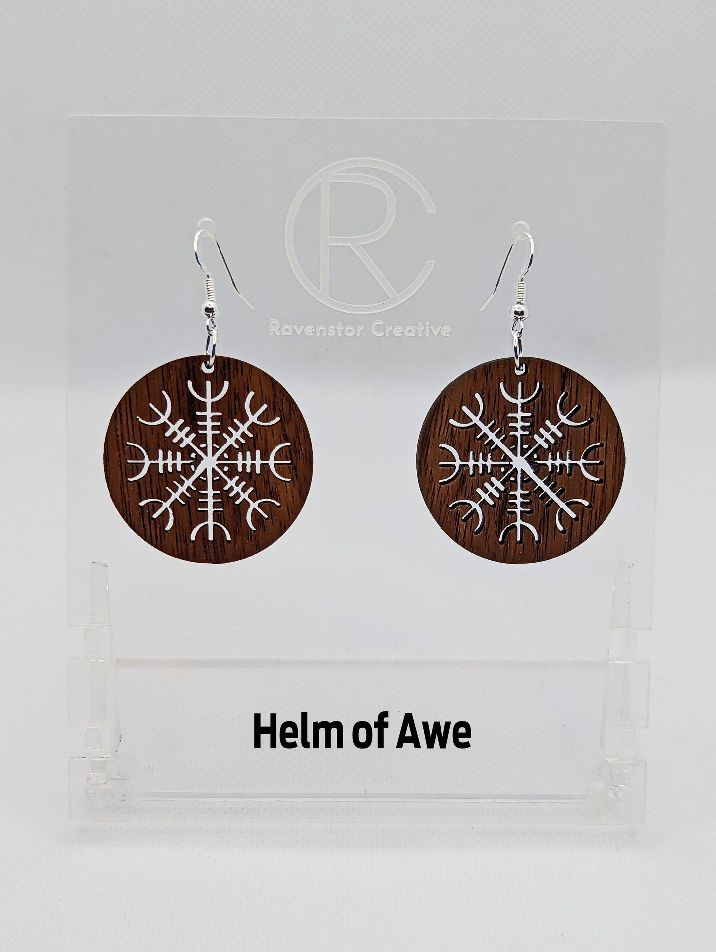 A pair of circle earrings with silver ear wire hook fixings. Cut out of the centre is the Helm of Awe Nordic charm. They are made from walnut wood. They are against a white background on a clear stand with the Ravenstor Creative logo on it.