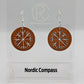 A pair of circle earrings with silver ear wire hook fixings. Cut out of the centre is the Nordic Compass charm. They are made from cherry wood. They are against a white background on a clear stand with the Ravenstor Creative logo on it.