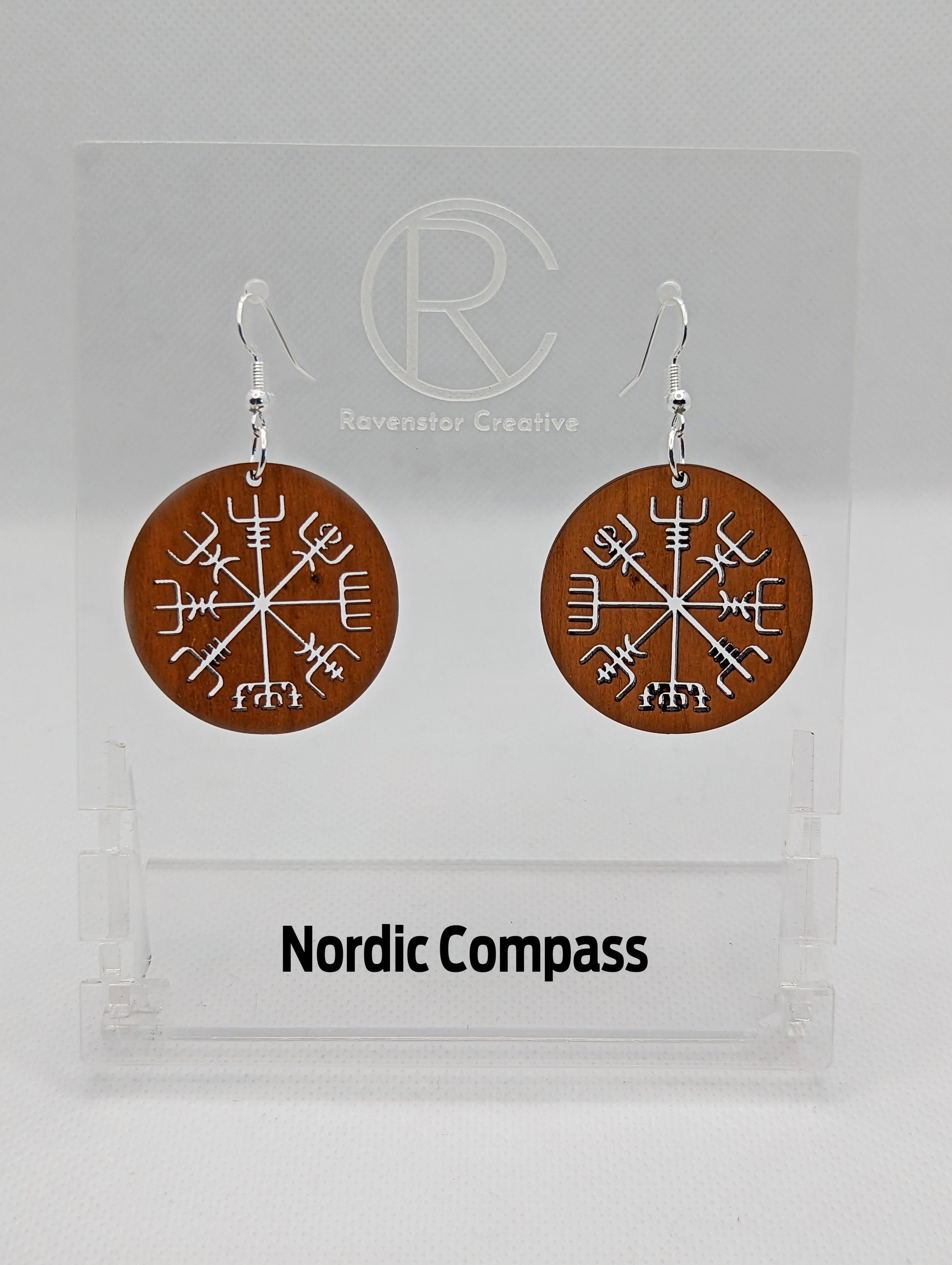 A pair of circle earrings with silver ear wire hook fixings. Cut out of the centre is the Nordic Compass charm. They are made from cherry wood. They are against a white background on a clear stand with the Ravenstor Creative logo on it.