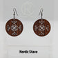 A pair of circle earrings with black ear wire hook fixings. Cut out of the centre is the Nordic Stave charm. They are made from walnut wood. They are against a white background on a clear stand with the Ravenstor Creative logo on it.