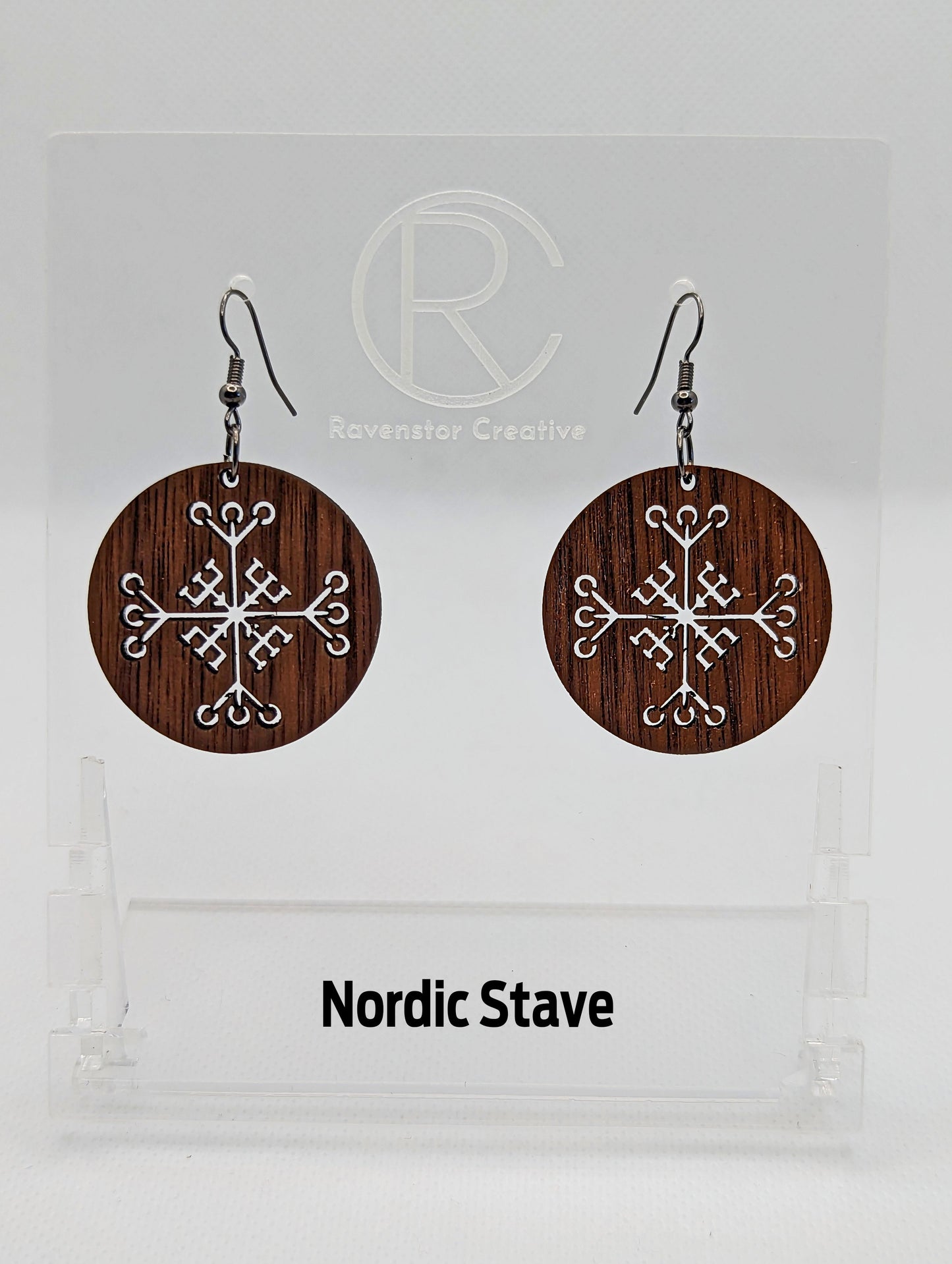 A pair of circle earrings with black ear wire hook fixings. Cut out of the centre is the Nordic Stave charm. They are made from walnut wood. They are against a white background on a clear stand with the Ravenstor Creative logo on it.