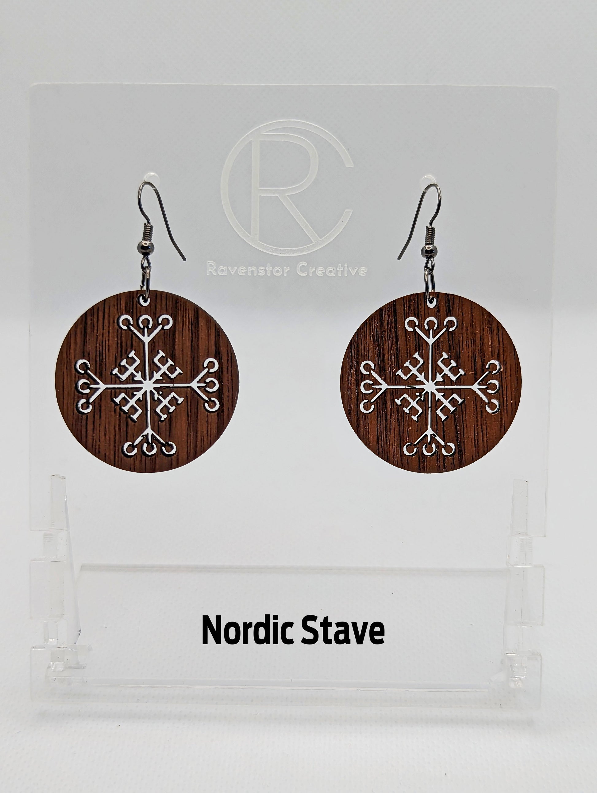 A pair of circle earrings with black ear wire hook fixings. Cut out of the centre is the Nordic Stave charm. They are made from walnut wood. They are against a white background on a clear stand with the Ravenstor Creative logo on it.