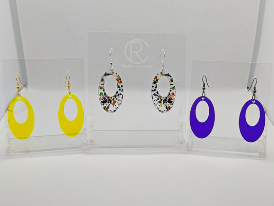 Shows three pairs of earrings that have ear wire hook fixings. They are oval shaped hoops. The colours are neon orange, lollipop glitter and purple. They are against a white background on see through stand with the ravenstor creative logo on.