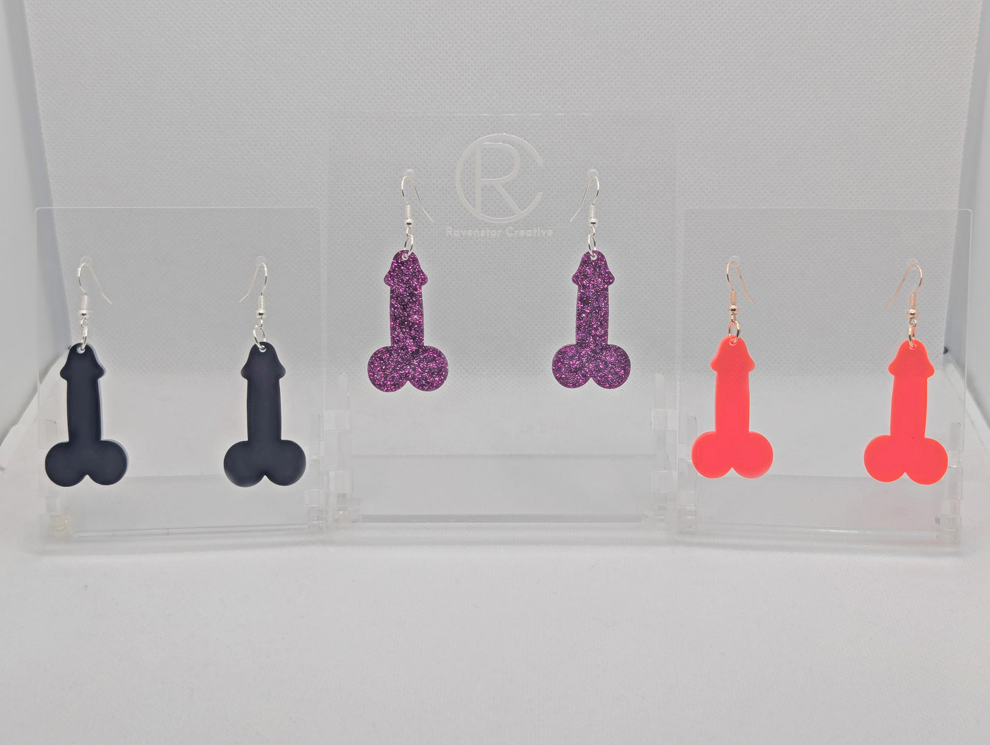 Image of three pairs of Earrings shaped like a Penis and Balls with ear wire hook fixings. These are ideal for funny Hen Dos or  Bachelorette parties. The colours are Black, Pink Glitter and Neon Pink. The are on Clear stands.