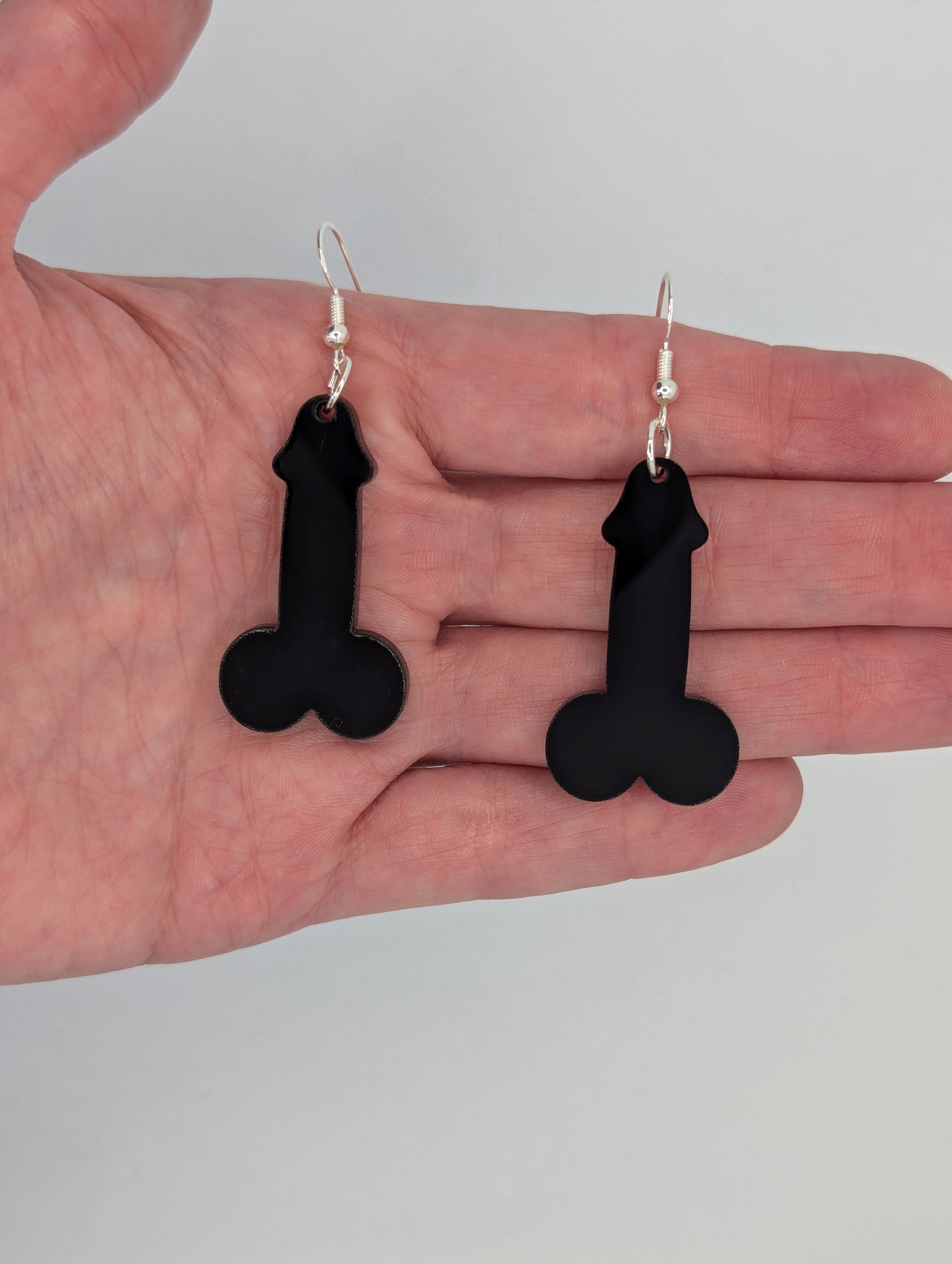 Image of a pair of earrings shaped like a Penis and Balls with Silver ear wire hook fixings. These are ideal for funny Hen Dos or Bachelorette parties. They are against a hand to show the size of two and a half finger widths in length and one wide