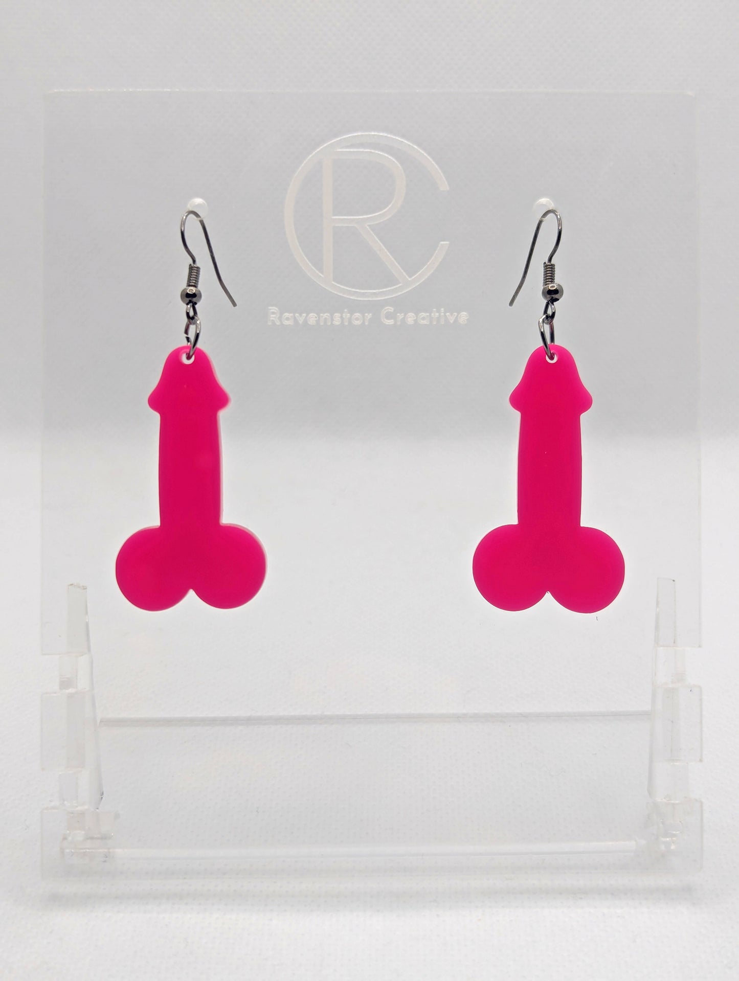 Image of a pair of earrings shaped like a Penis and Balls with Black wire hook fixings. These are ideal for funny Hen Dos or Bachelorette parties. The colour is Hot Pink and they are on clear stands against a white background.