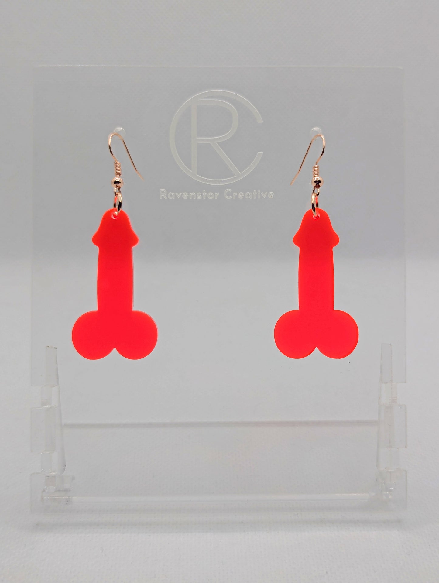 Image of a pair of earrings shaped like a Penis and Balls with Rose Gold ear wire hook fixings. These are ideal for funny Hen Dos or Bachelorette parties. The colour is Neon Pink and they are on clear stands against a white background.