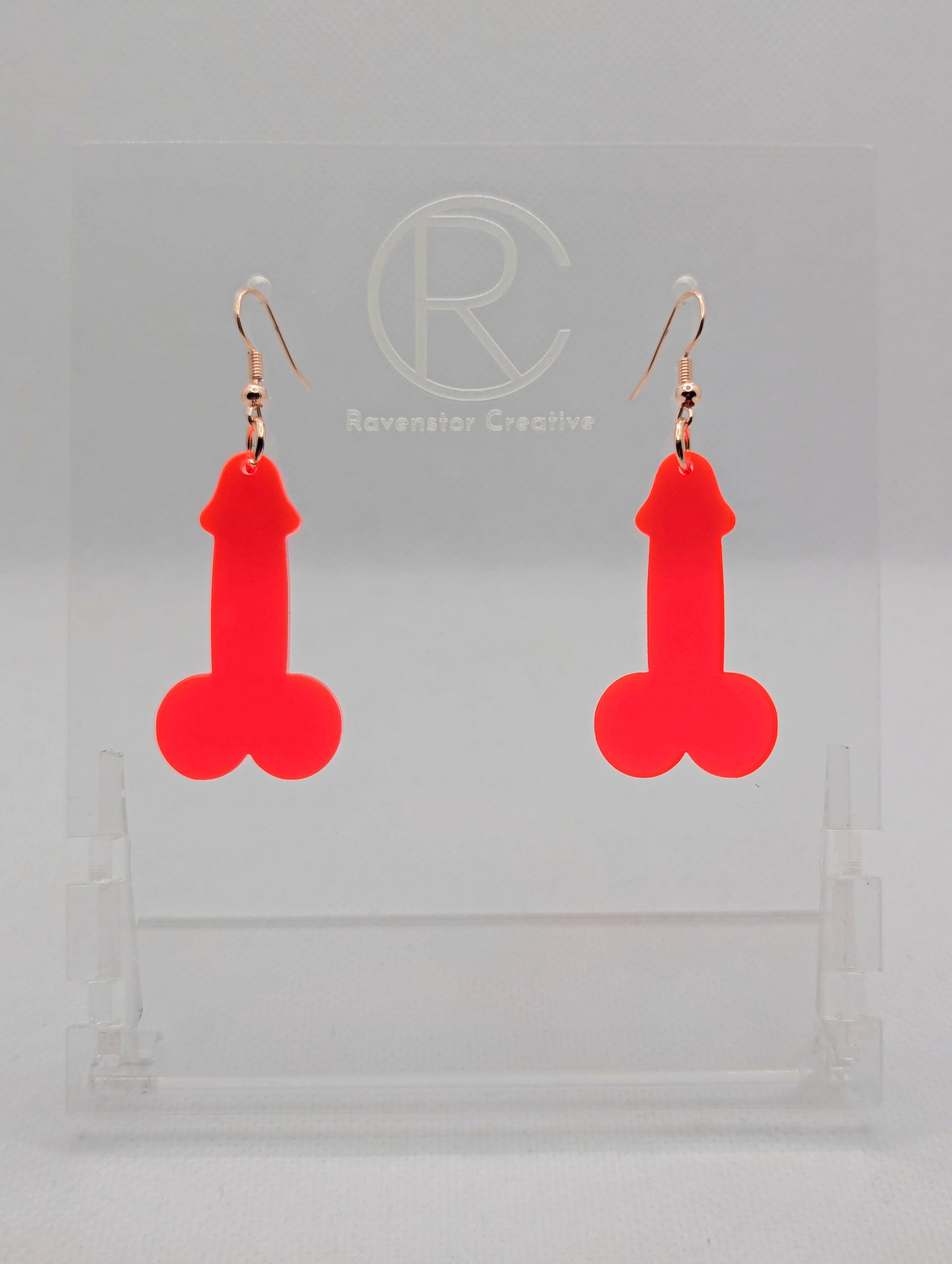 Image of a pair of earrings shaped like a Penis and Balls with Rose Gold ear wire hook fixings. These are ideal for funny Hen Dos or Bachelorette parties. The colour is Neon Pink and they are on clear stands against a white background.