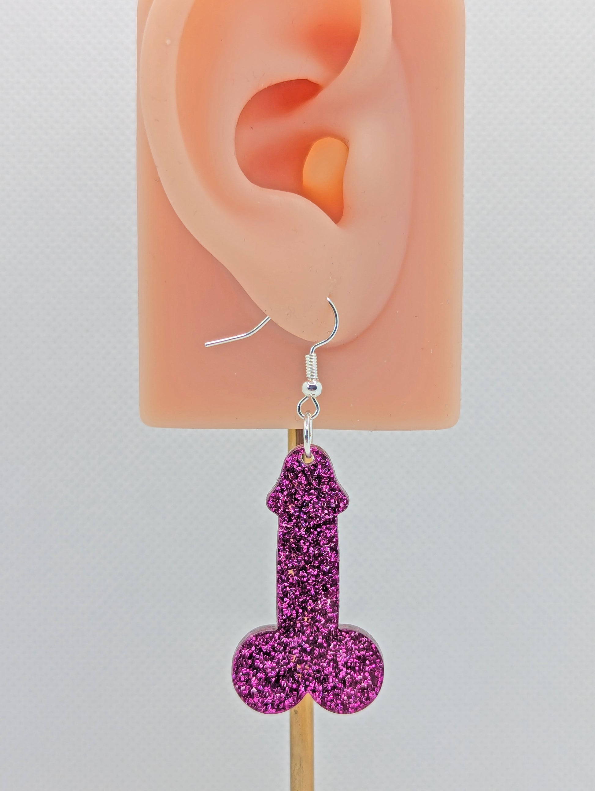 Image of an earring shaped like a Penis and Balls with a Silver ear wire hook fixing. These are ideal for funny Hen Dos or  Bachelorette parties. The colour is Pink Glitter and is on an ear to show the size.