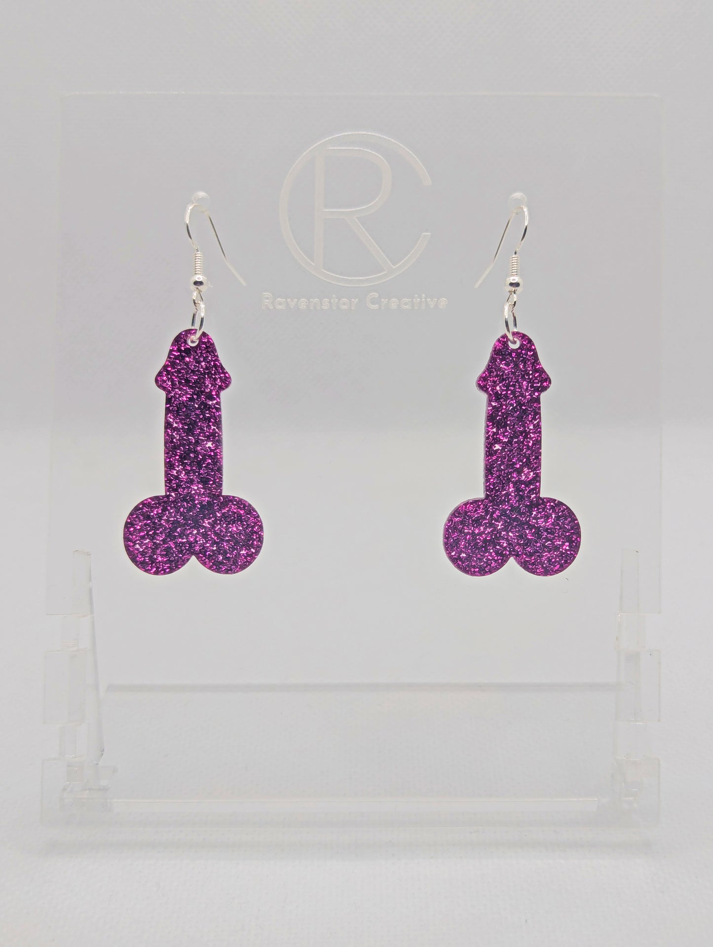 Image of a pair of earrings shaped like a Penis and Balls with Silver ear wire hook fixings. These are ideal for funny Hen Dos or Bachelorette parties. The colour is Pink Glitter and they are on clear stands against a white background.