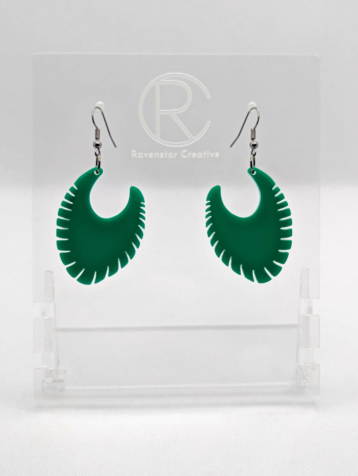 Image of a pair of acrylic earrings with black ear wire hook fixings. They are Ribbed half moon shapes with ridges around one side. They are Green and are on clear stands with the Ravenstor Creative logo on them against a white background.