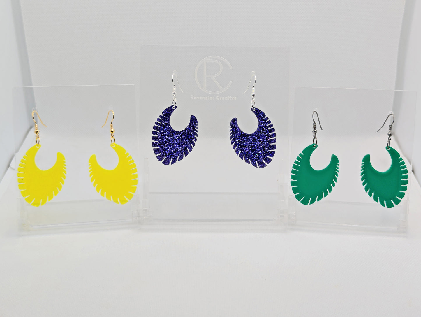 Image of three pairs of earrings with ear wire hook fixings. They are Ribbed half moon shapes with ridges around one side. The colours are Neon Orange, Purple Glitter and Green. They are on clear stands against a white background.