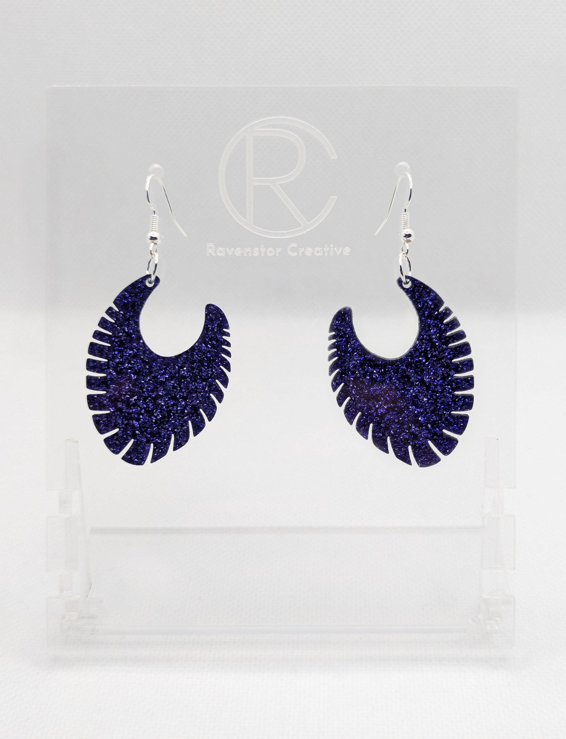 Image of a pair of acrylic earrings with Silver ear wire hook fixings. They are Ribbed half moon shapes with ridges around one side. They are Purple Glitter and are on clear stands with the Ravenstor Creative logo on them against a white background.