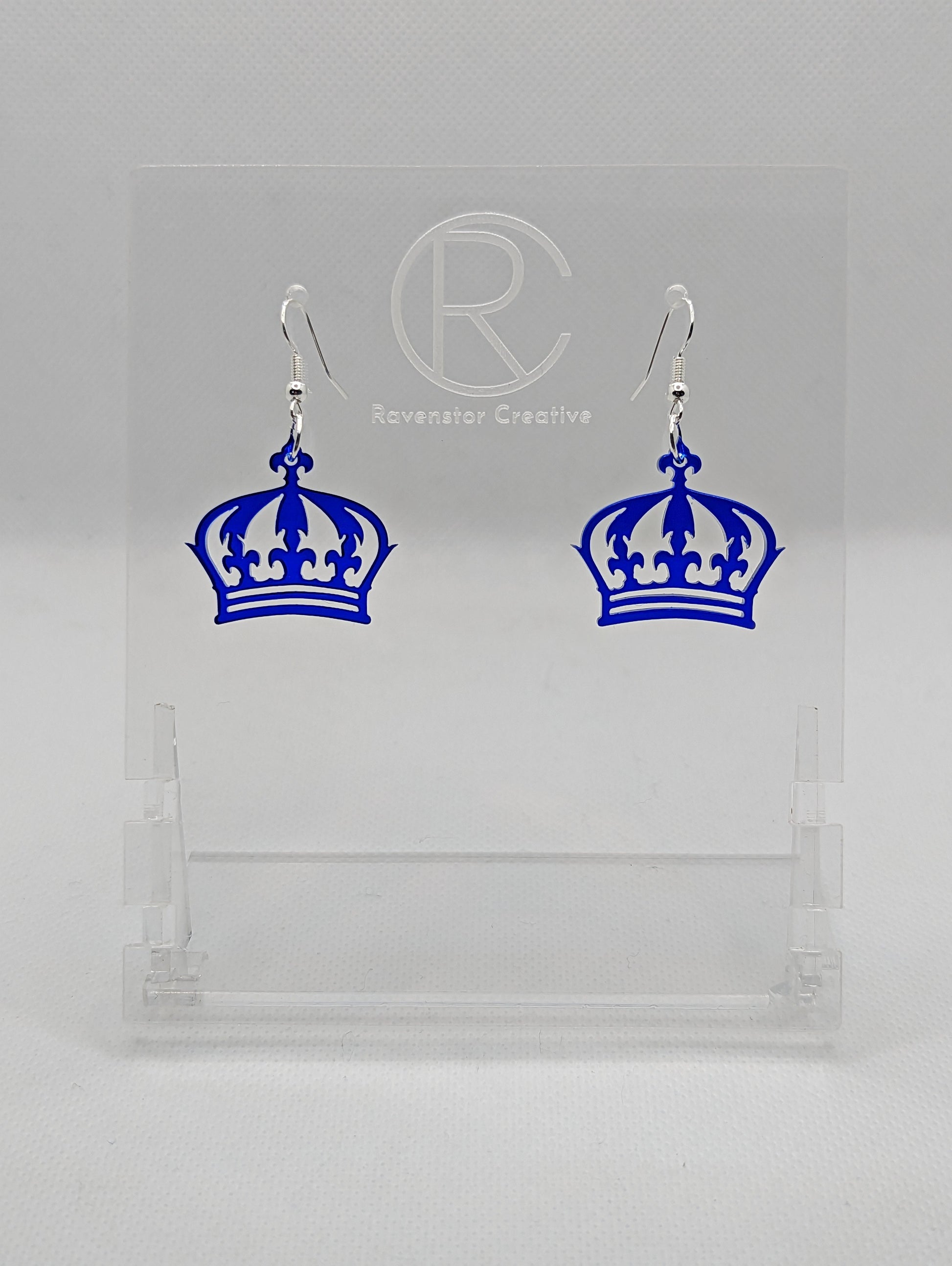 Shows a pair of acrylic earrings with silver ear wire hook fixings. They are Royal Crown shapes. The colour is Royal Blue Glitter. They are against a white background on clear stands with the Ravenstor Creative logo