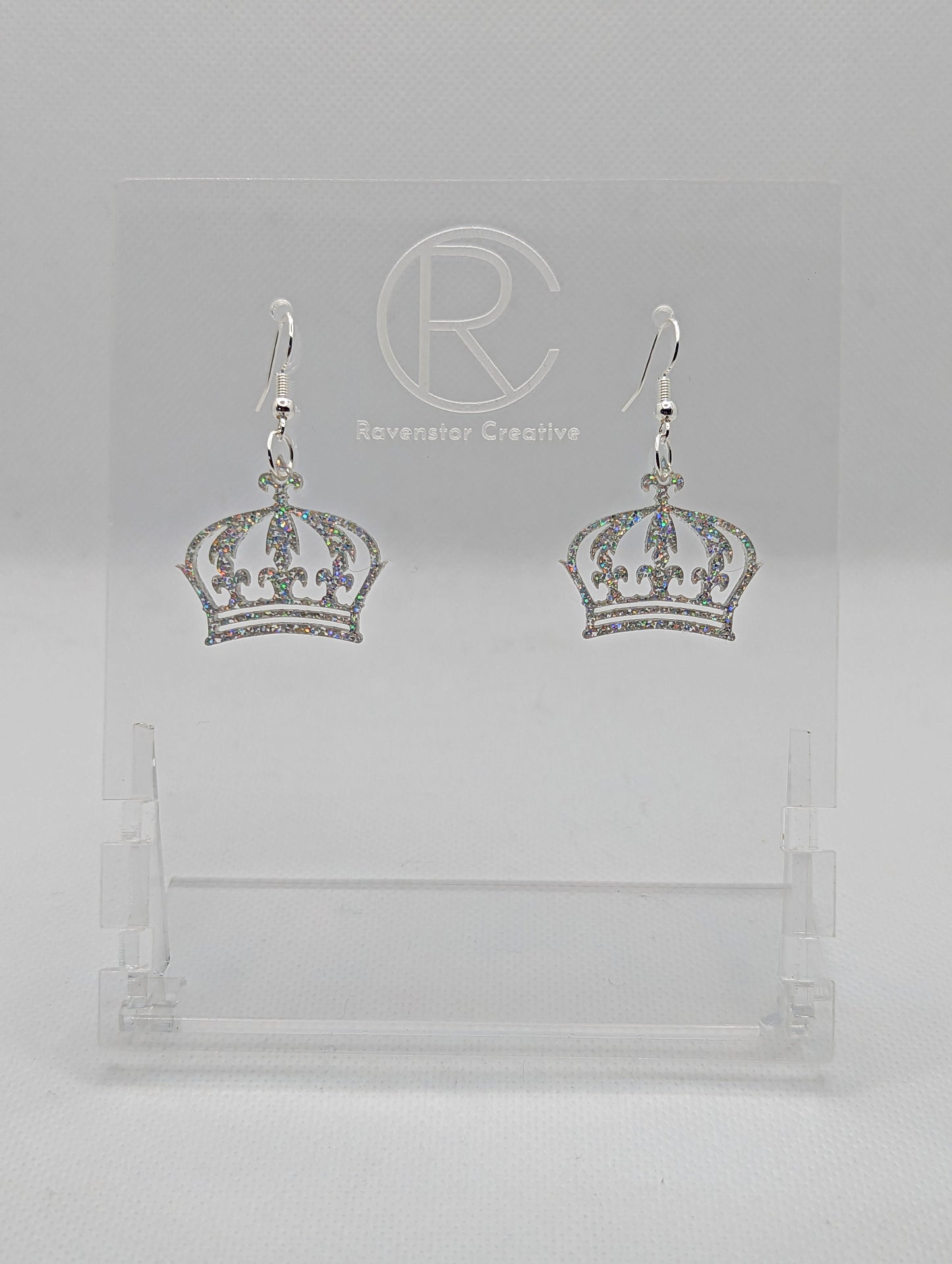 Shows a pair of acrylic earrings with silver ear wire hook fixings. They are Royal Crown shapes. The colour is Holographic Glitter. They are against a white background on clear stands with the Ravenstor Creative logo.