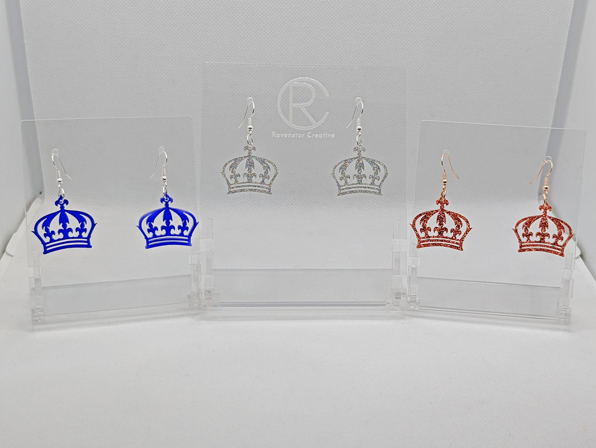 Three pairs of acrylic earrings with ear wire hook fixings. They are Royal Crown shapes. The colours are Translucent Blue, Holographic Glitter and Copper Glitter. They are against a white background on clear stands with the Ravenstor Creative logo.