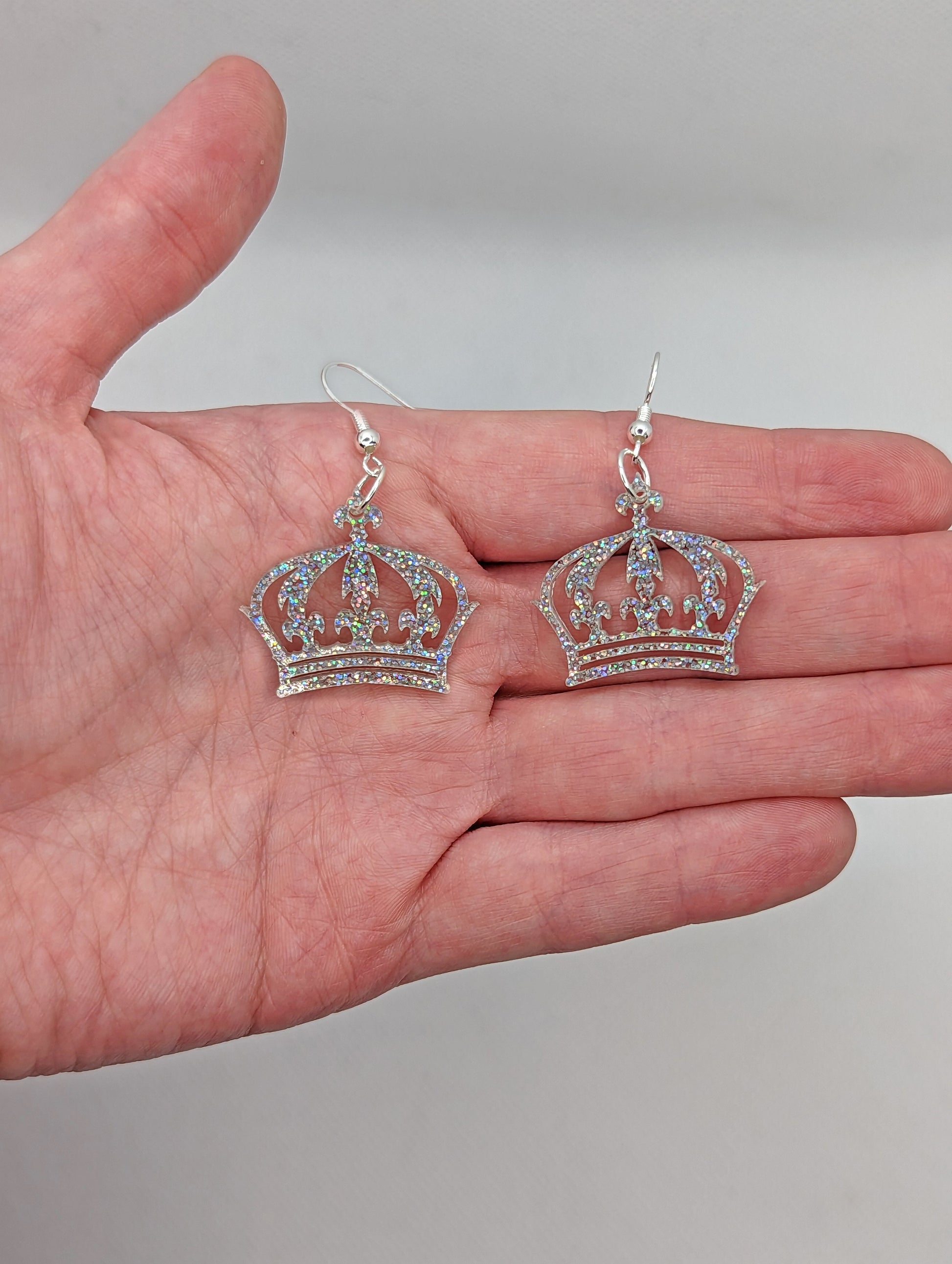 Shows a pair of acrylic earrings with silver ear wire hook fixings. They are Royal Crown shapes. It is against a hand showing the size of one and a half finger widths length and same again wide. They are Holographic Glitter colour.