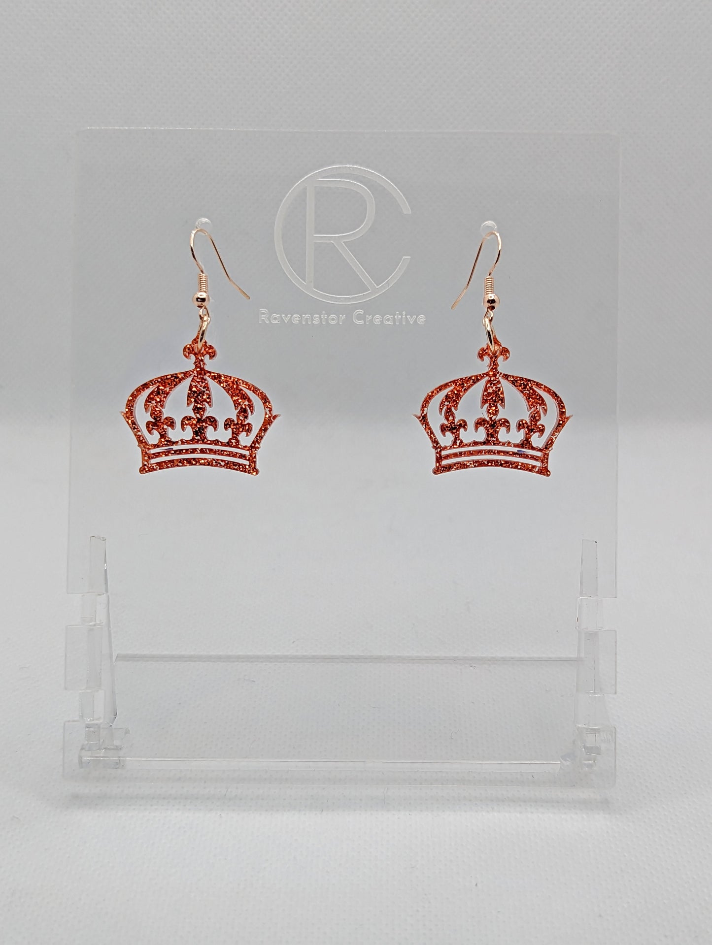 Shows a pair of acrylic earrings with Rose Gold ear wire hook fixings. They are Royal Crown shapes. The colour is Copper Glitter. They are against a white background on clear stands with the Ravenstor Creative logo.