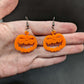 Image of a pair of scary Halloween pumpkin shaped earrings on a hand showing the size against a dark background, they are one and a half finger widths length and two wide. They are orange and have silver fixings.