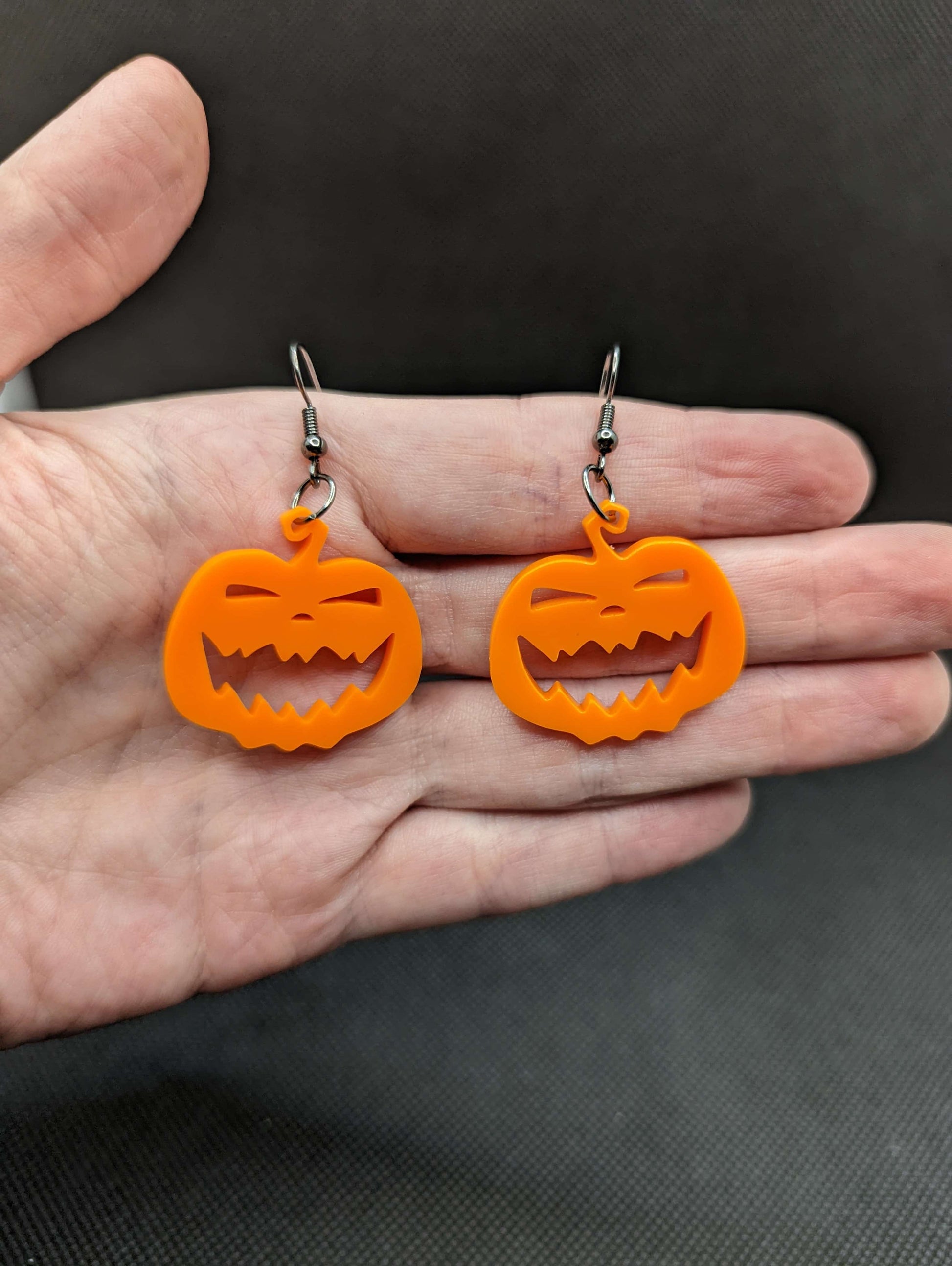 Image of a pair of scary Halloween pumpkin shaped earrings on a hand showing the size against a dark background, they are one and a half finger widths length and two wide. They are orange and have silver fixings.