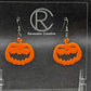 Shows a pair of earrings with black ear wire hook fixings. They are Scary Halloween pumpkin shapes. The colour of them are orange. They are on a see through stand that has the Ravenstor Creative Logo and its against a dark background.