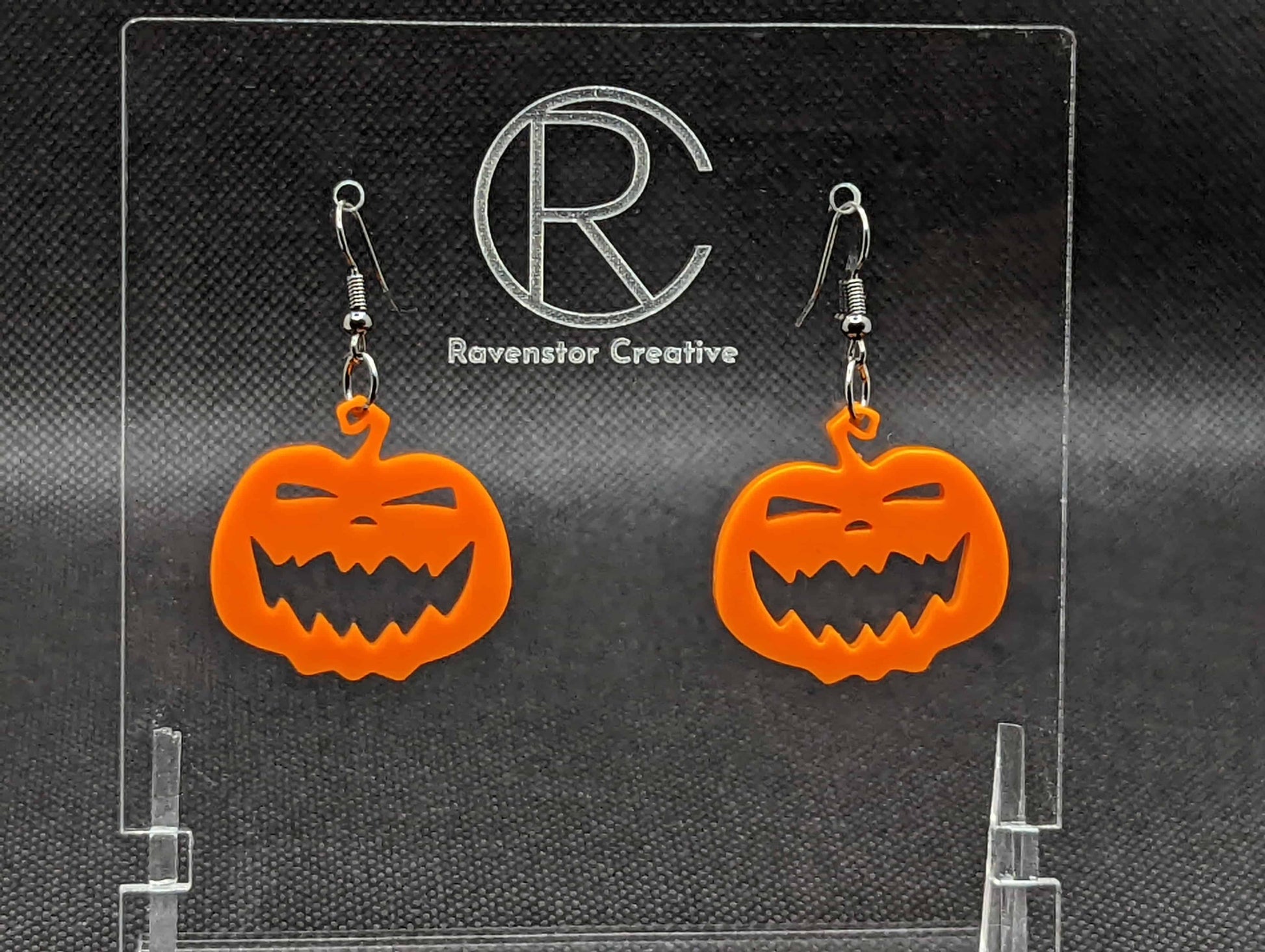 Shows a pair of earrings with black ear wire hook fixings. They are Scary Halloween pumpkin shapes. The colour of them are orange. They are on a see through stand that has the Ravenstor Creative Logo and its against a dark background.