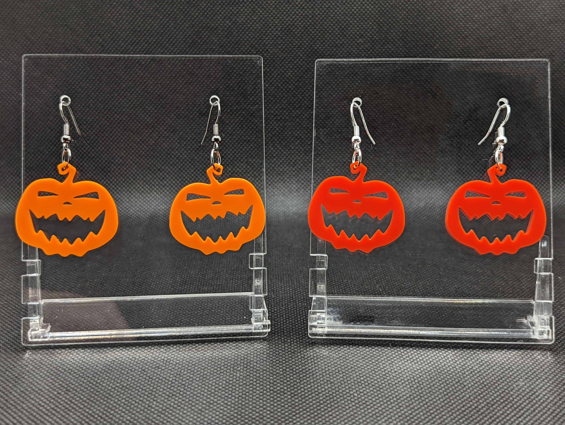 Shows 2 pairs of earrings with ear wire hook fixings. They are scary Halloween pumpkin shapes. The colours of them are Orange and Red. They are on see through stands with the Ravenstor Creative logo on them against a dark background.