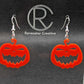 Shows a pair of earrings with silver ear wire hook fixings. They are Scary Halloween pumpkin shapes. The colour of them are red. They are on a see through stand that has the Ravenstor Creative Logo and its against a dark background.