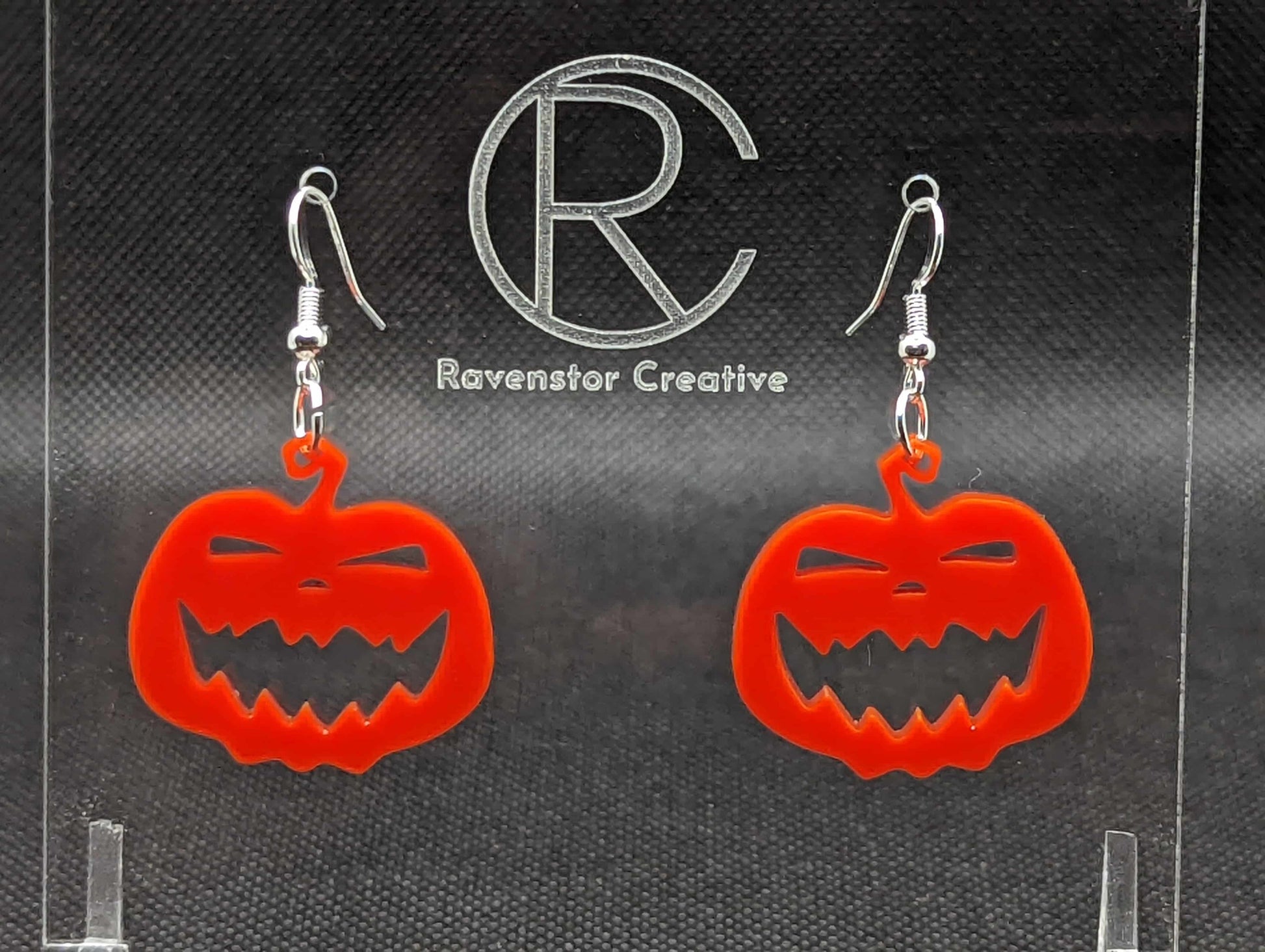 Shows a pair of earrings with silver ear wire hook fixings. They are Scary Halloween pumpkin shapes. The colour of them are red. They are on a see through stand that has the Ravenstor Creative Logo and its against a dark background.