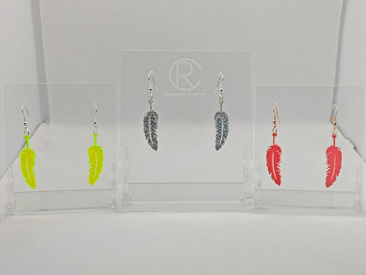 Shows three pairs of earrings that have ear wire hook fixings. They are pretty feather shaped. The colours are neon green, silver glitter and sherbet pink. They are against a white background on see through stand with the Ravenstor Creative logo on.